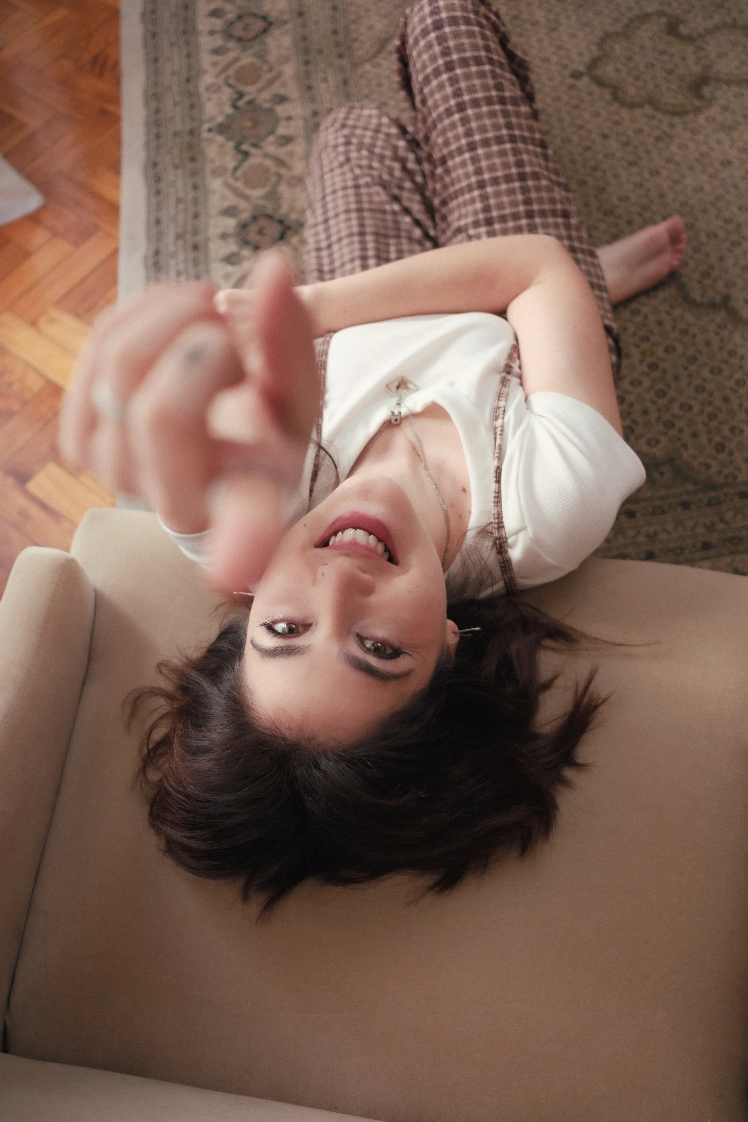Joven mujer de cabello oscuro y suelto está acostada en un sofá, vista desde arriba, sonriendo y extendiendo la mano hacia la cámara.
