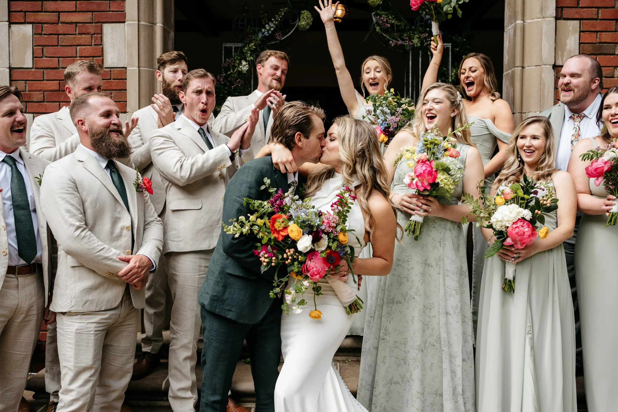 A wedding celebration with a bride and groom kissing, surrounded by bridesmaids and groomsmen, with some cheering and raising their hands outside a brick building.