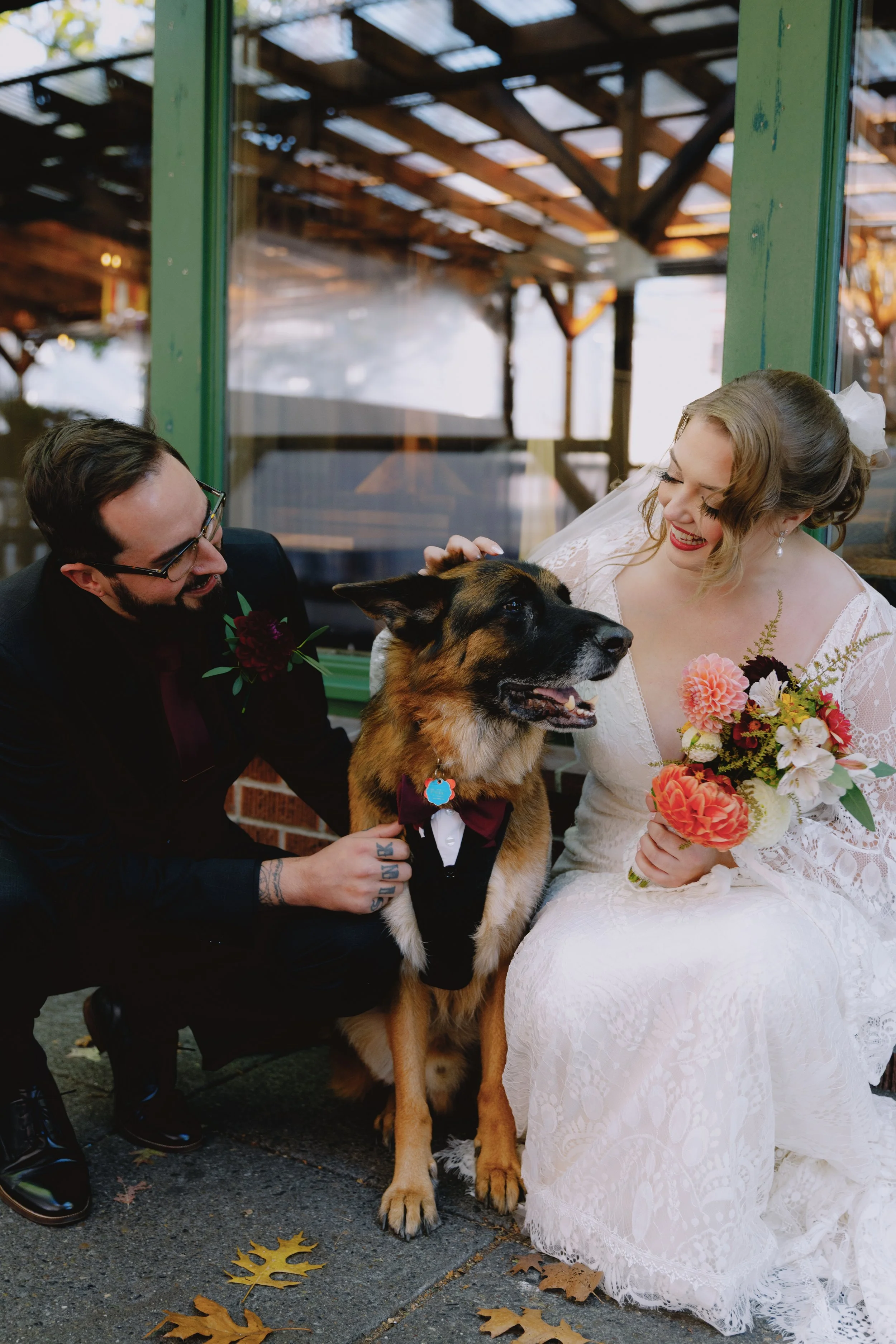 A bride and groom with their dog, dressed in a tuxedo, during their wedding celebration outdoors. The bride is holding a bouquet, and both are smiling and interacting with the dog.