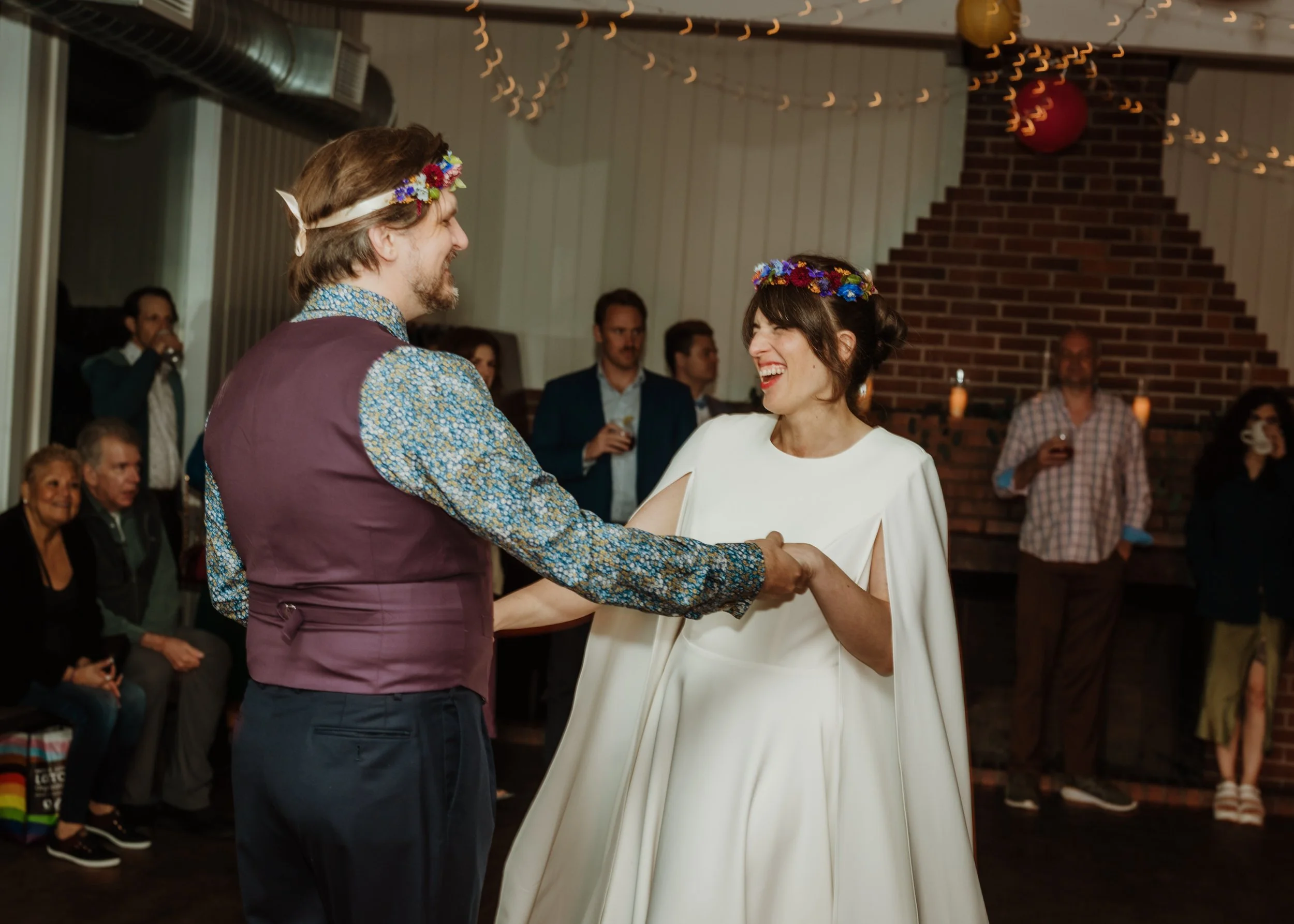 A couple dancing at a wedding, holding hands and smiling, with guests watching in the background. They are wearing flower crowns.