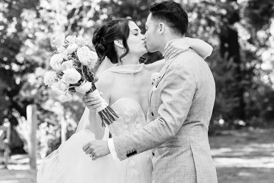 A wedding ceremony outdoors with a couple exchanging vows under a white arch decorated with flowers, surrounded by seated guests in bright attire, with green trees in the background.