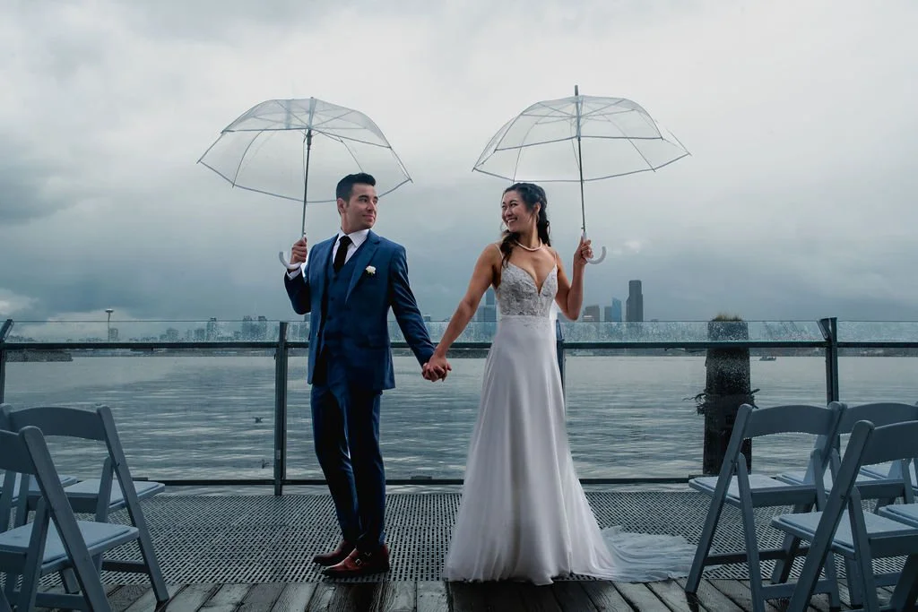 A newlywed couple holding hands and umbrellas on a rainy day by the waterfront, with city buildings in the background.