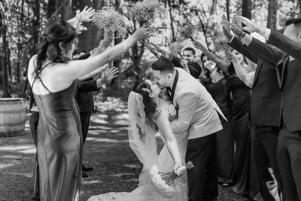 Couple kissing during wedding celebration with friends and family in a wooded outdoor setting.