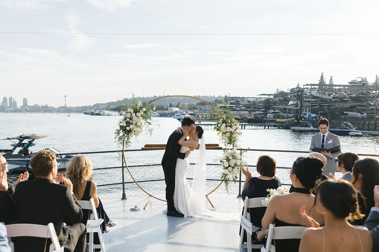 A bride and groom kissing during a wedding ceremony by the water, with guests watching and clapping, and a city skyline in the background.