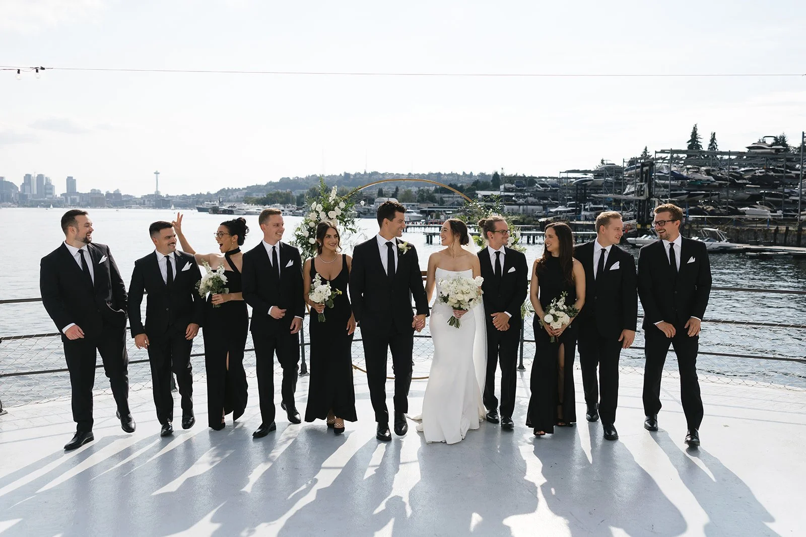 A wedding party of ten, dressed in black and white formal attire, standing on a dock with water and city skyline in the background. The bride and groom are holding hands in the center, surrounded by bridesmaids and groomsmen holding bouquets and smil