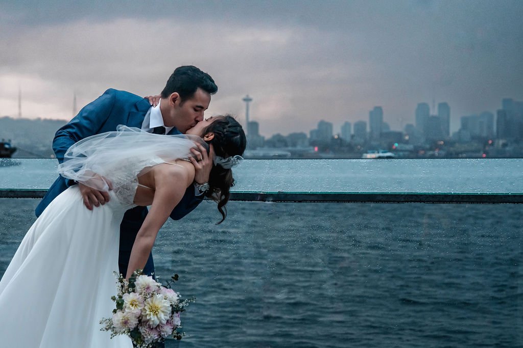 A newlywed couple sharing a kiss on a rooftop overlooking a city skyline with water in the foreground during overcast weather.