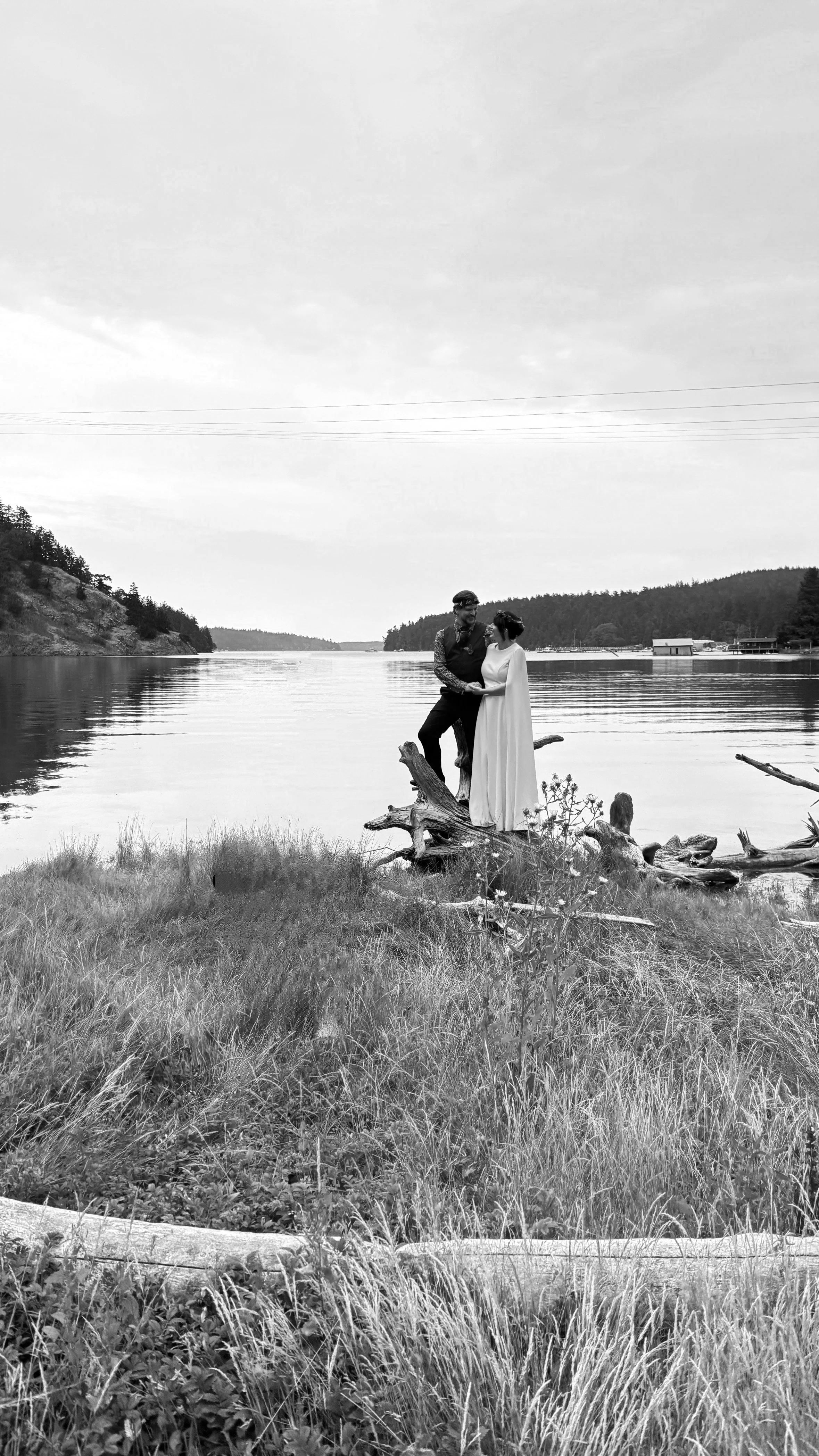 A couple standing on a fallen tree by the water, with the man in dark clothing and the woman in a white dress, holding hands, near a lakeshore with mountains in the background.