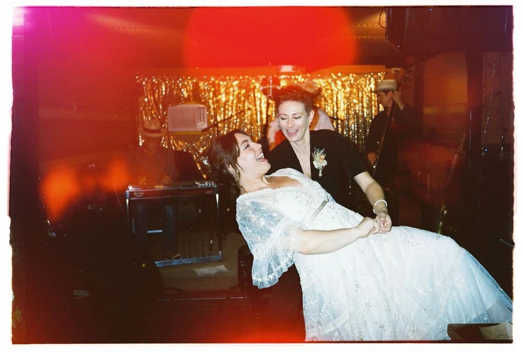 Two women dancing and smiling at a celebration, one in a white dress and the other in black, with a sparkly gold backdrop and musicians in the background.