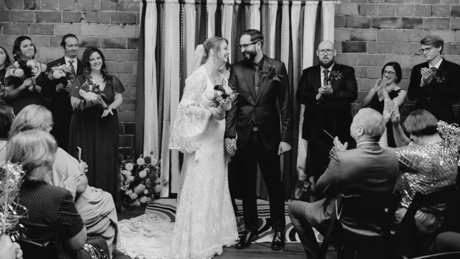 A couple dancing closely at an indoor event with a crowd watching, brick walls, and colorful lighting.