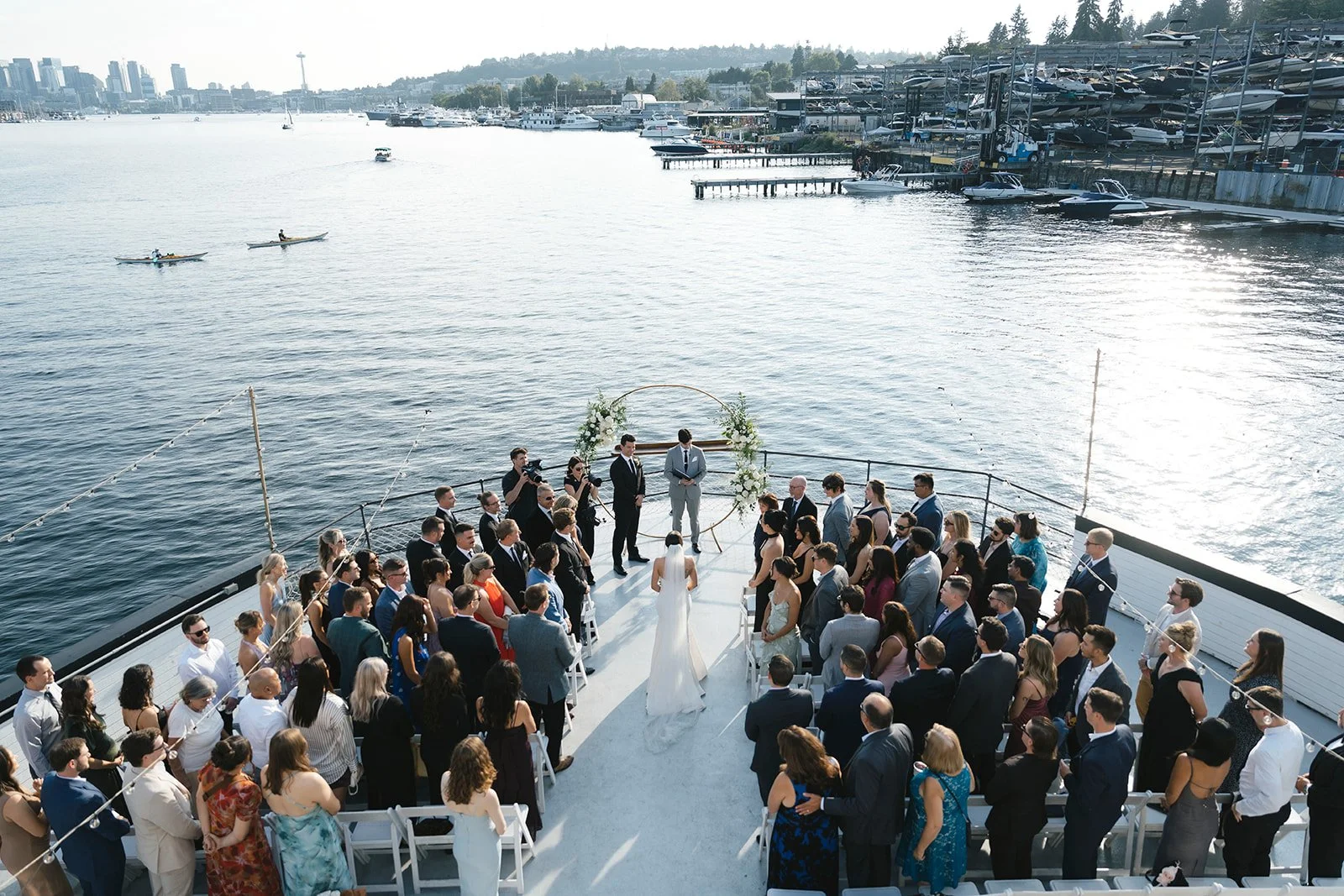 Wedding ceremony on a boat deck by the water with many guests, a bride and groom at the altar, and a city skyline in the background.
