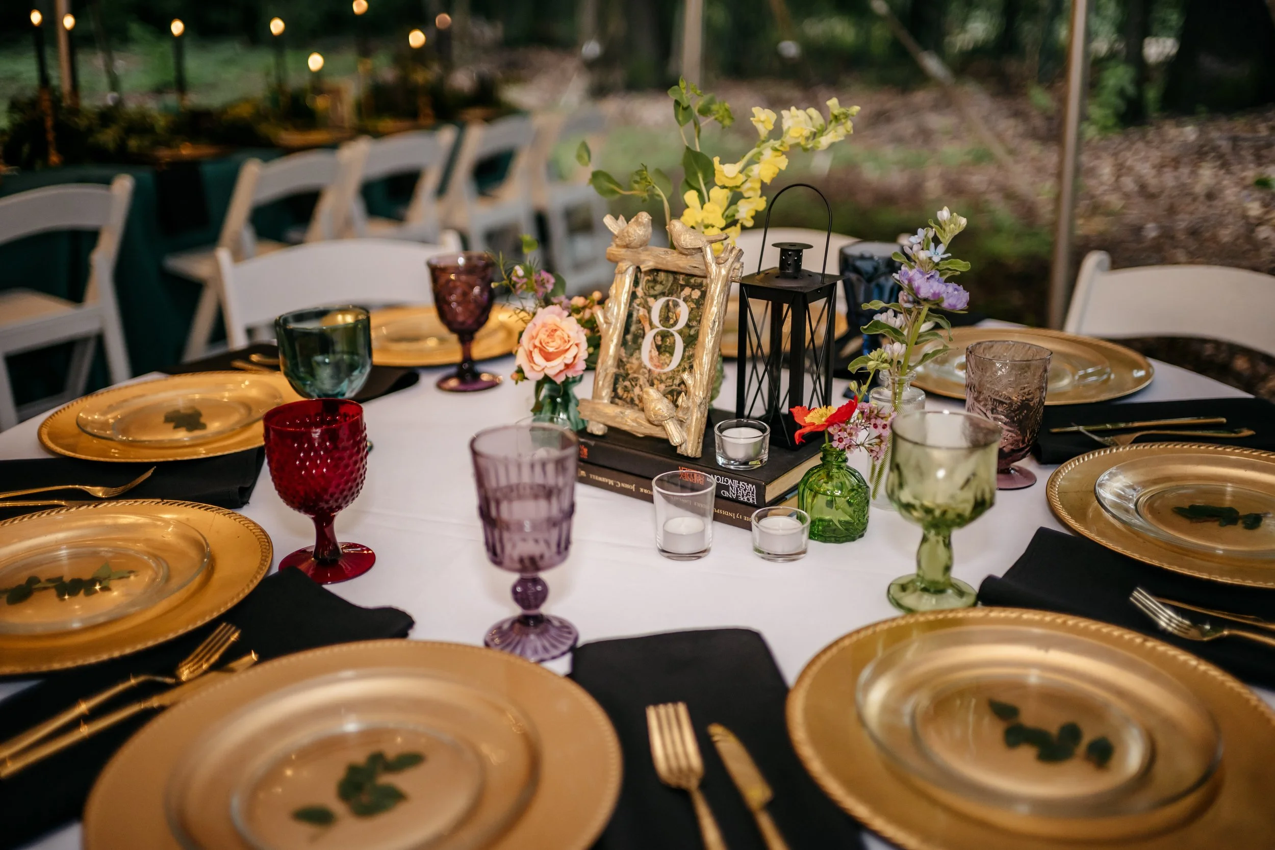 Elegant outdoor dining table with gold charger plates, colorful goblets, black napkins, and floral centerpieces in vases, set for a celebration.