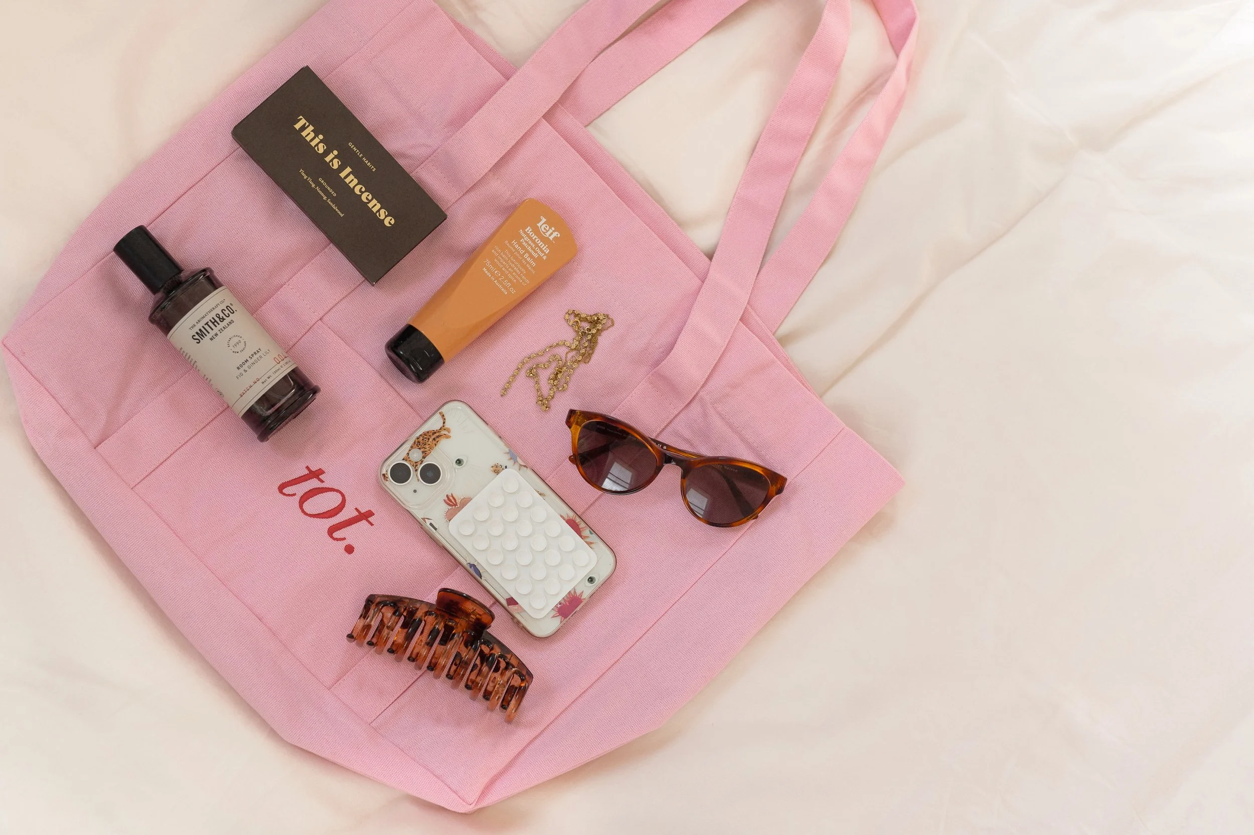 Pink tote bag with the words 'tot.' in red, containing a bottle of perfume, a tube of hand cream, a gold chain necklace, sunglasses, a phone with a patterned case, a hair clip, and a card that reads 'This is Incense' on a cream-colored surface.