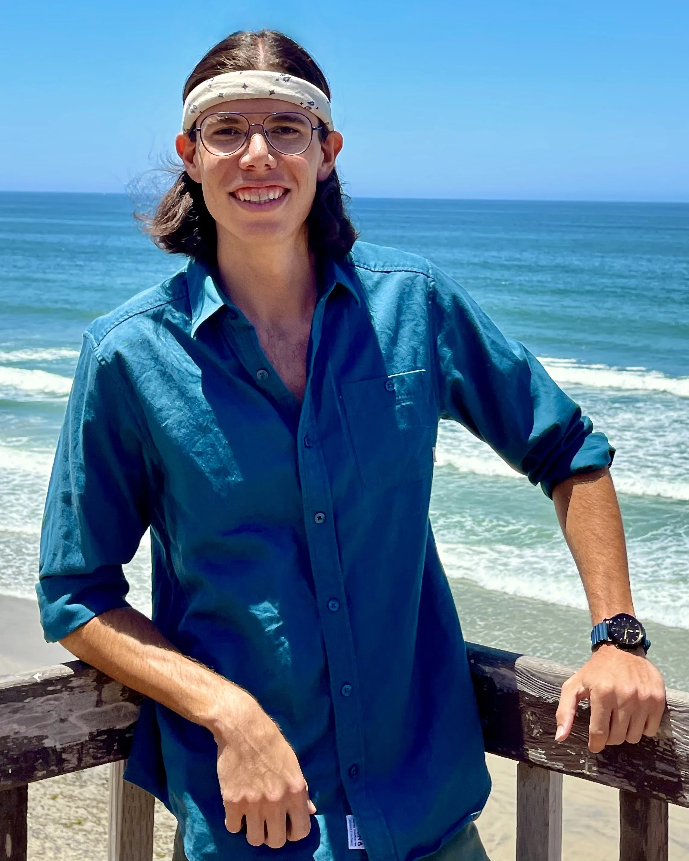 A man with long brown hair, wearing a white bandana, glasses, and a blue shirt, standing on a beach with the ocean and clear sky in the background.