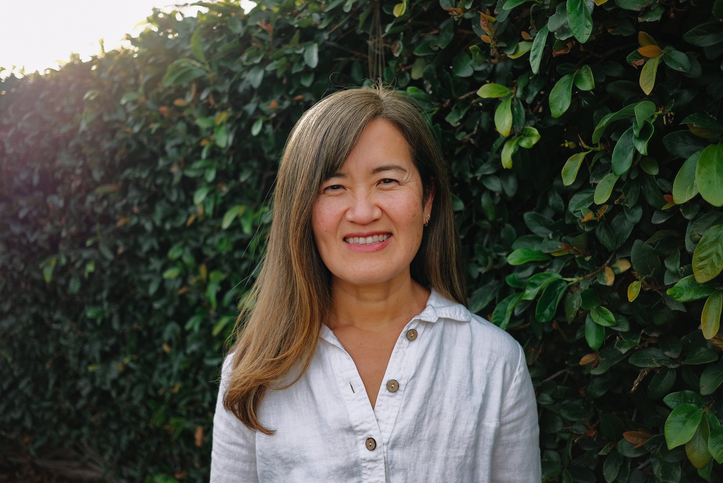 A woman with long brown hair smiling in front of a leafy hedge, wearing a white button-up shirt.