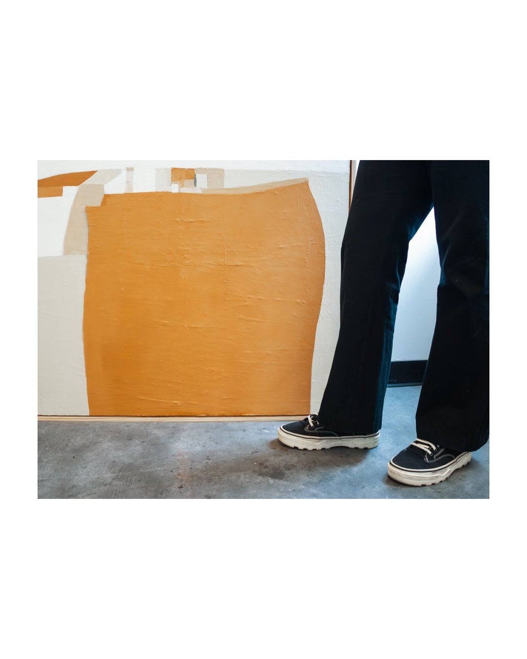 The painting’s intricate texture and geometric details are highlighted, with a glimpse of the artist’s legs and high-top sneakers holding the artwork, offering a sense of scale and process.