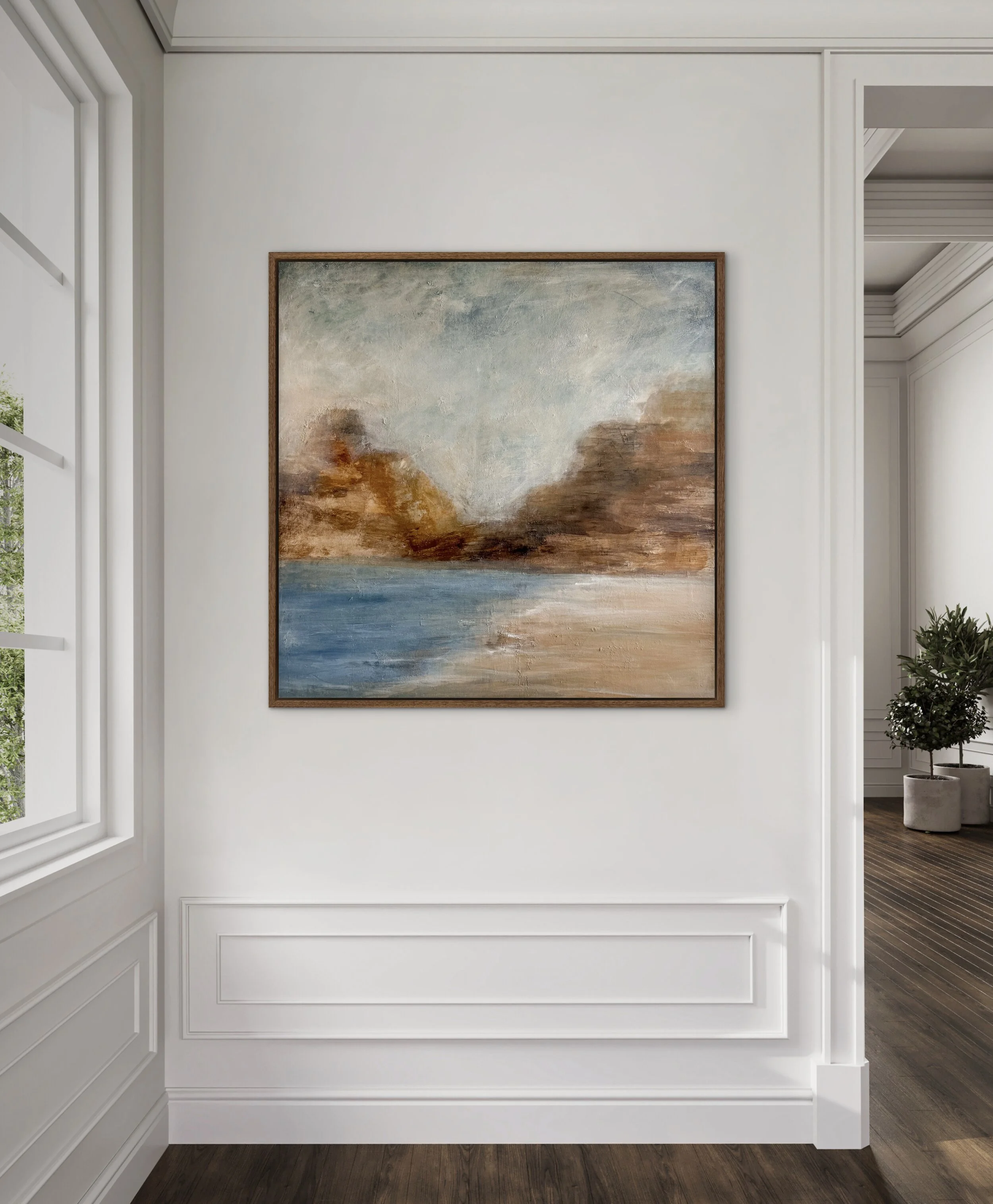 Large framed abstract landscape painting with soft blue water, sandy beige shoreline, earthy rust-colored cliffs, and a pale sky, displayed on a white paneled wall in a refined, light-filled interior.