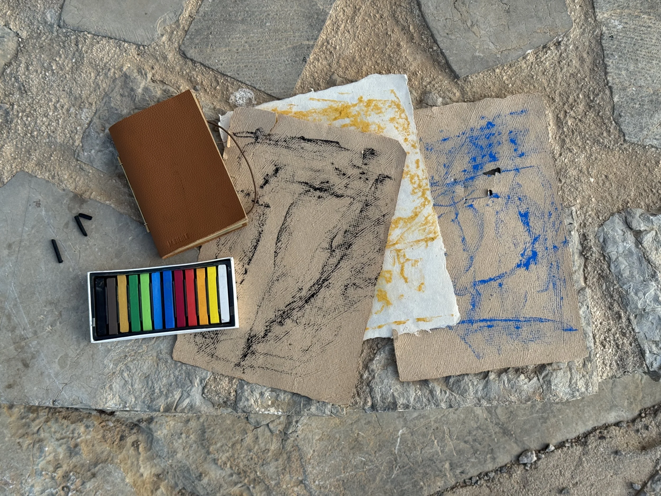 Charcoal and pastel experiments on handmade papers arranged on a stone bench during an artist immersion in Hydra, Greece.