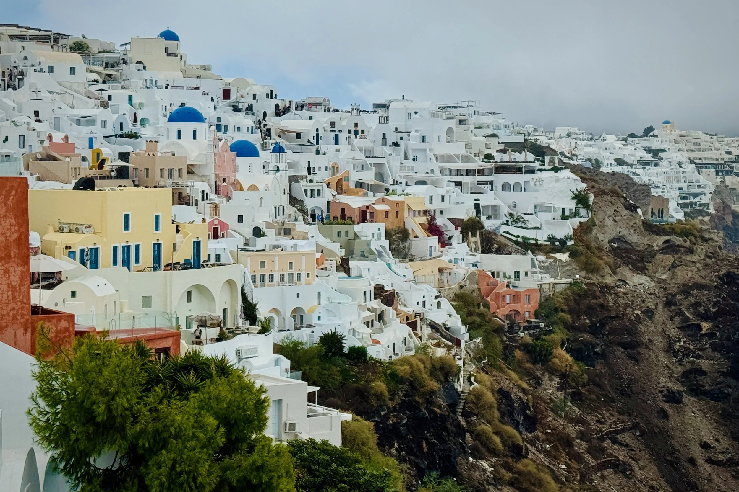View across Oia in Santorini, Greece, showing white Cycladic buildings, blue domes, colorful facades, and volcanic cliffs.