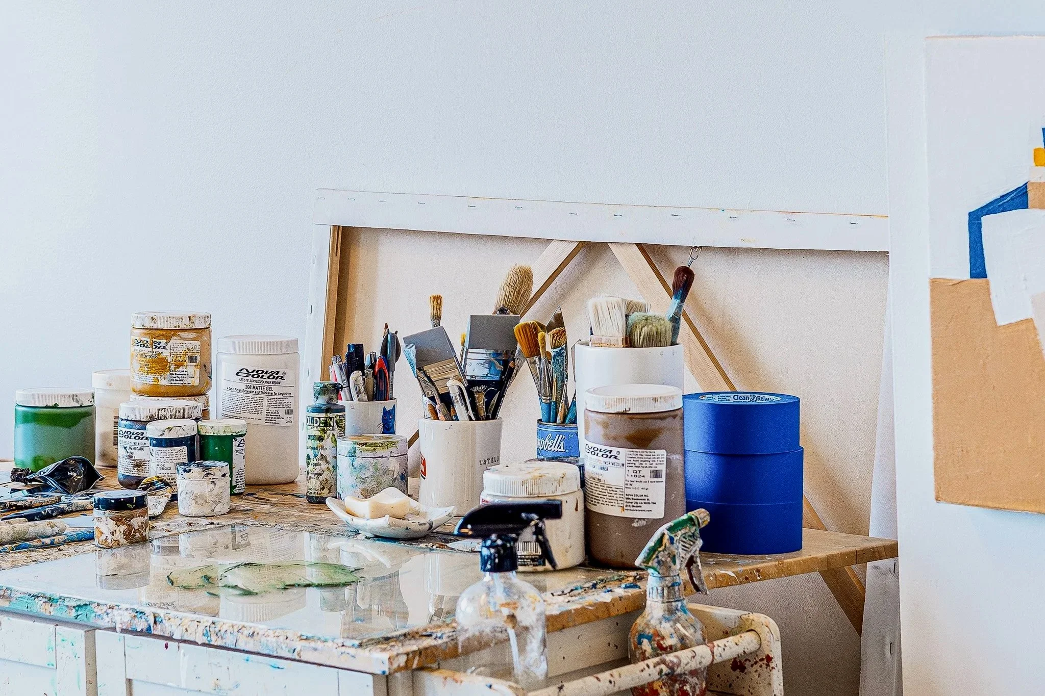 Artist’s studio worktable with paint jars, brushes, tools, and materials arranged on a well-used surface, showing an in-progress creative workspace.