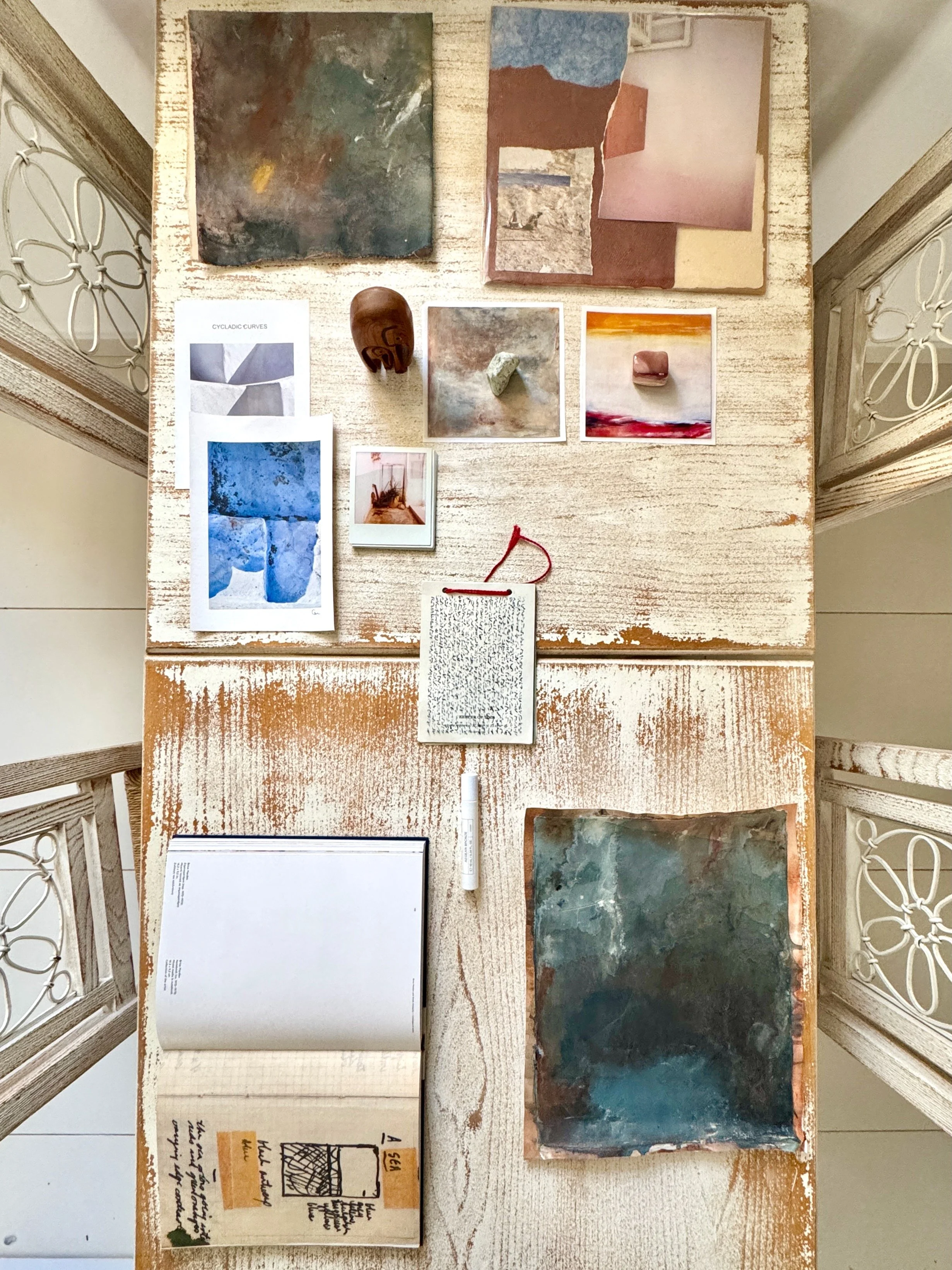 Artist inspiration table in Hydra, Greece, with a small abstract painting, photographs, found objects, sketchbook, and personal artifacts arranged for a creative immersion.
