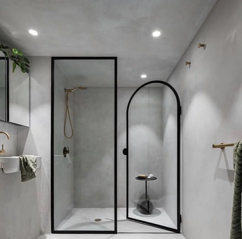 Modern bathroom with a walk-in shower, black framing, white walls, ceiling-mounted lights, a small sink with a gold faucet, and a towel on a gold towel rack.
