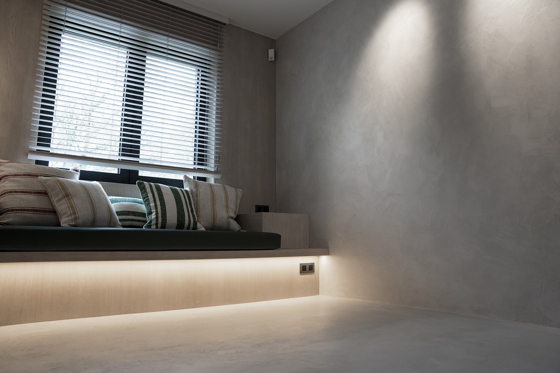 Minimalist interior with a bed built into a raised platform under a large window with horizontal blinds, decorated with striped and solid pillows, and illuminated with soft under-bed lighting.