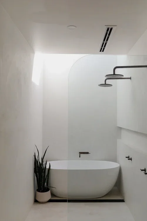 Minimalist bathroom with a large freestanding bathtub, a tall green plant in a white pot, modern shower with rain showerhead, and white walls and ceiling.