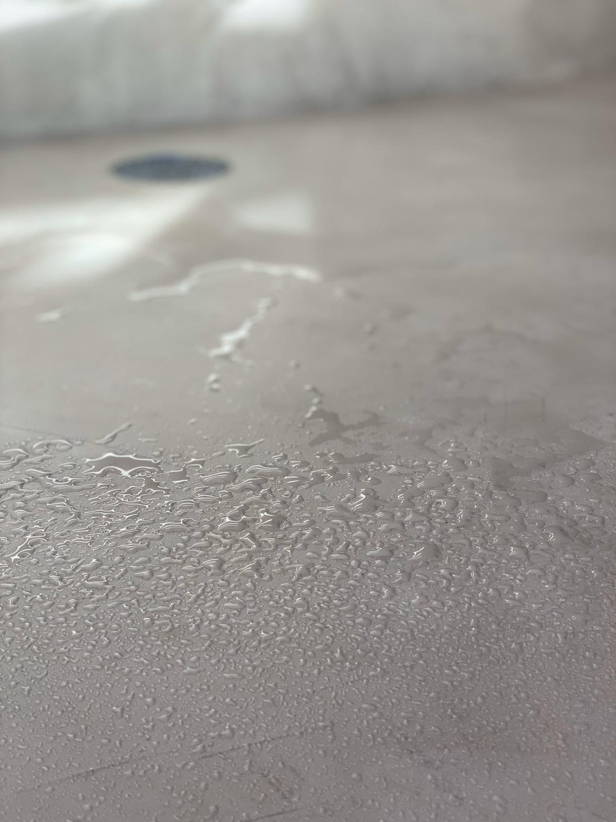 Close-up of a wet surface with water droplets and small puddles, possibly a metallic or smooth surface, with a drain visible in the background.
