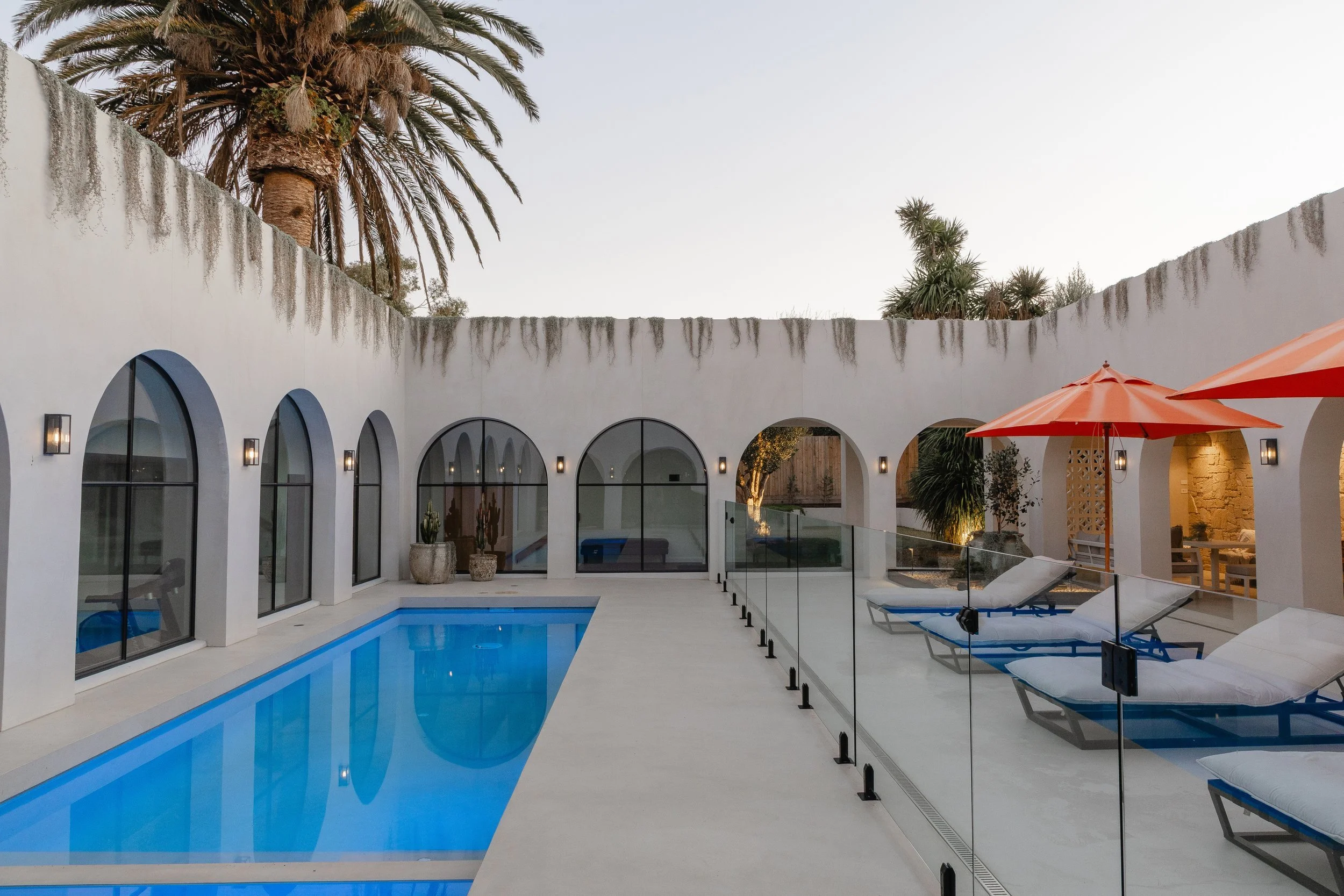A modern private pool area with lounge chairs and large orange umbrellas, surrounded by a white wall with arched windows and palm trees in the background.