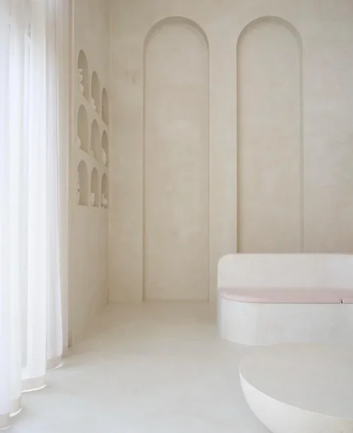 Minimalist beige interior with built-in niche shelves and a bench, featuring tall arched wall details and sheer curtains on the left side.