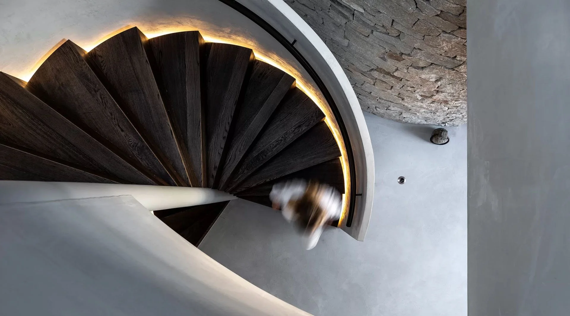 A top-down view of a curved wooden staircase with warm lighting beneath the steps, adjacent to a white wall and a textured brick wall, with a person blurred moving on the staircase.