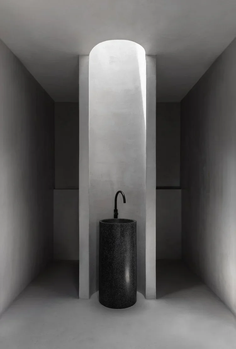 Minimalist space with a black cylindrical sink and a curved faucet, set against gray walls and a ceiling with a rounded opening letting in natural light.