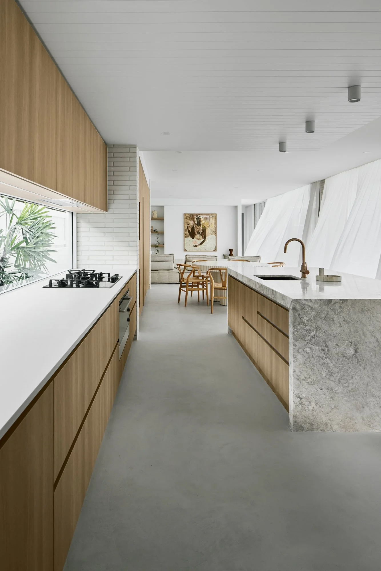 Modern kitchen with white countertops, wooden cabinets, gray stone island, and a view of the dining and living area with wooden chairs and artwork.
