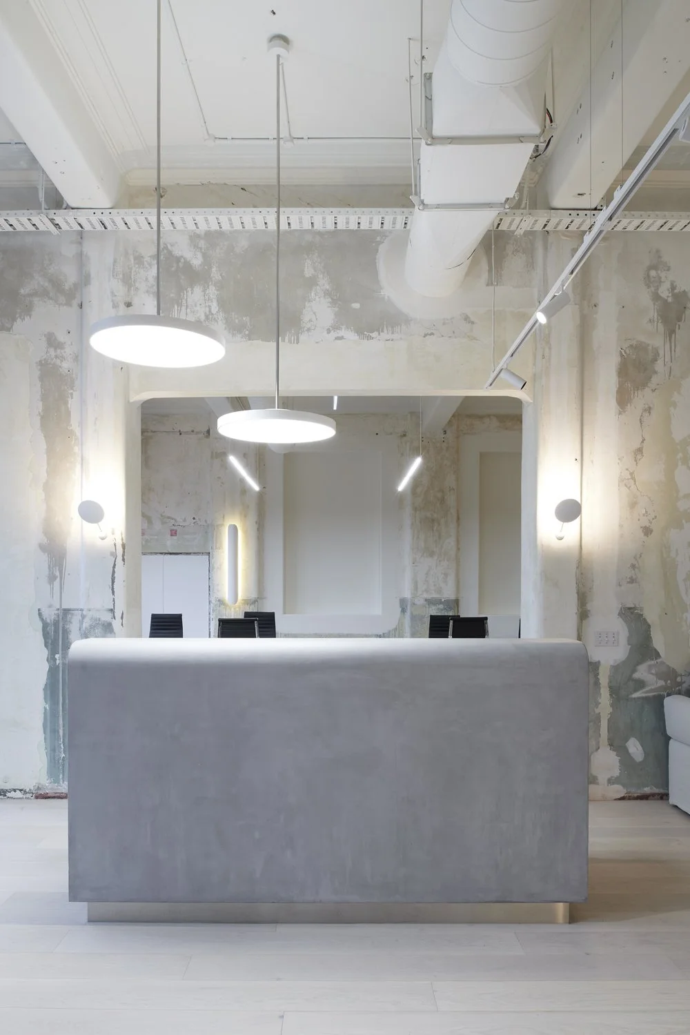 Modern office space with minimalist design, featuring a concrete front desk, black chairs, and contemporary lighting, with exposed ceiling pipes and unfinished walls.