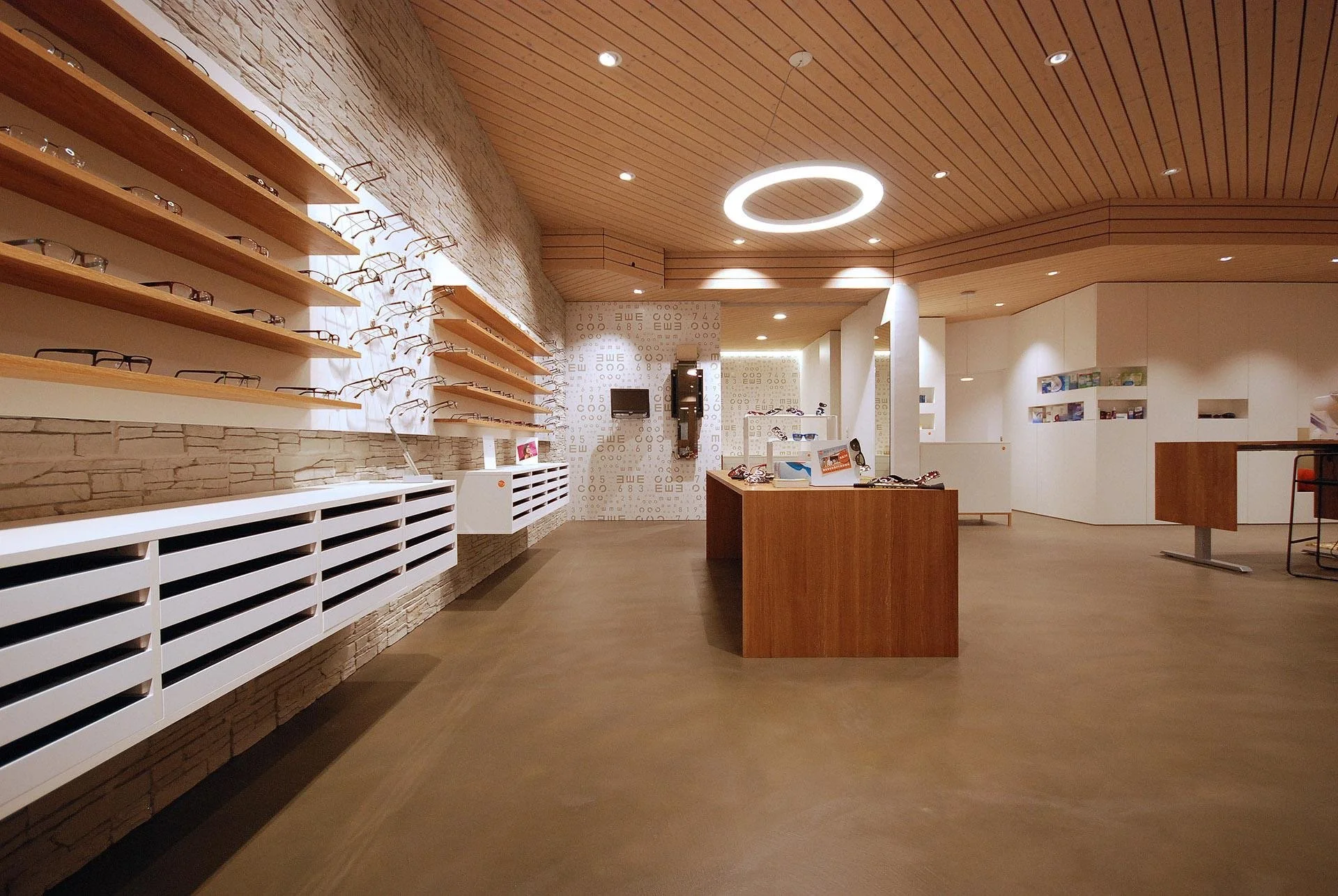 Inside an eyeglasses store with wooden and white display shelves, showing multiple pairs of glasses, a central wooden table with eyewear, and a monitor mounted on a patterned wall.