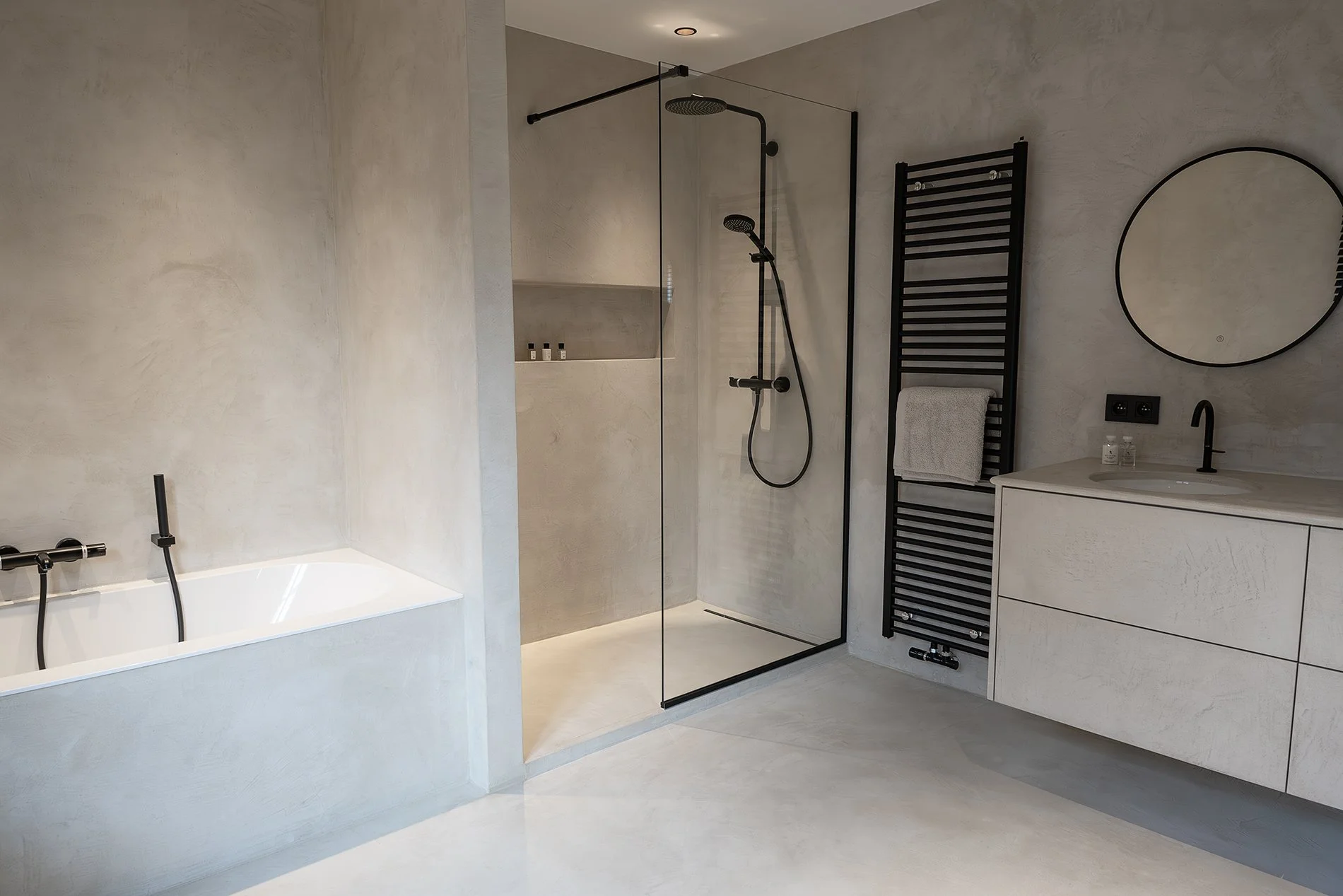 Modern minimalist bathroom with a bathtub, glass-enclosed shower, black towel heater, vanity with round mirror, sink, and toiletries.