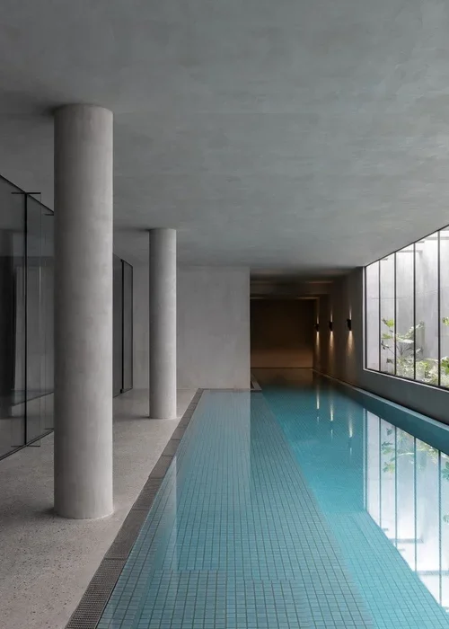 Indoor swimming pool with large floor-to-ceiling windows, concrete columns, and modern decor.