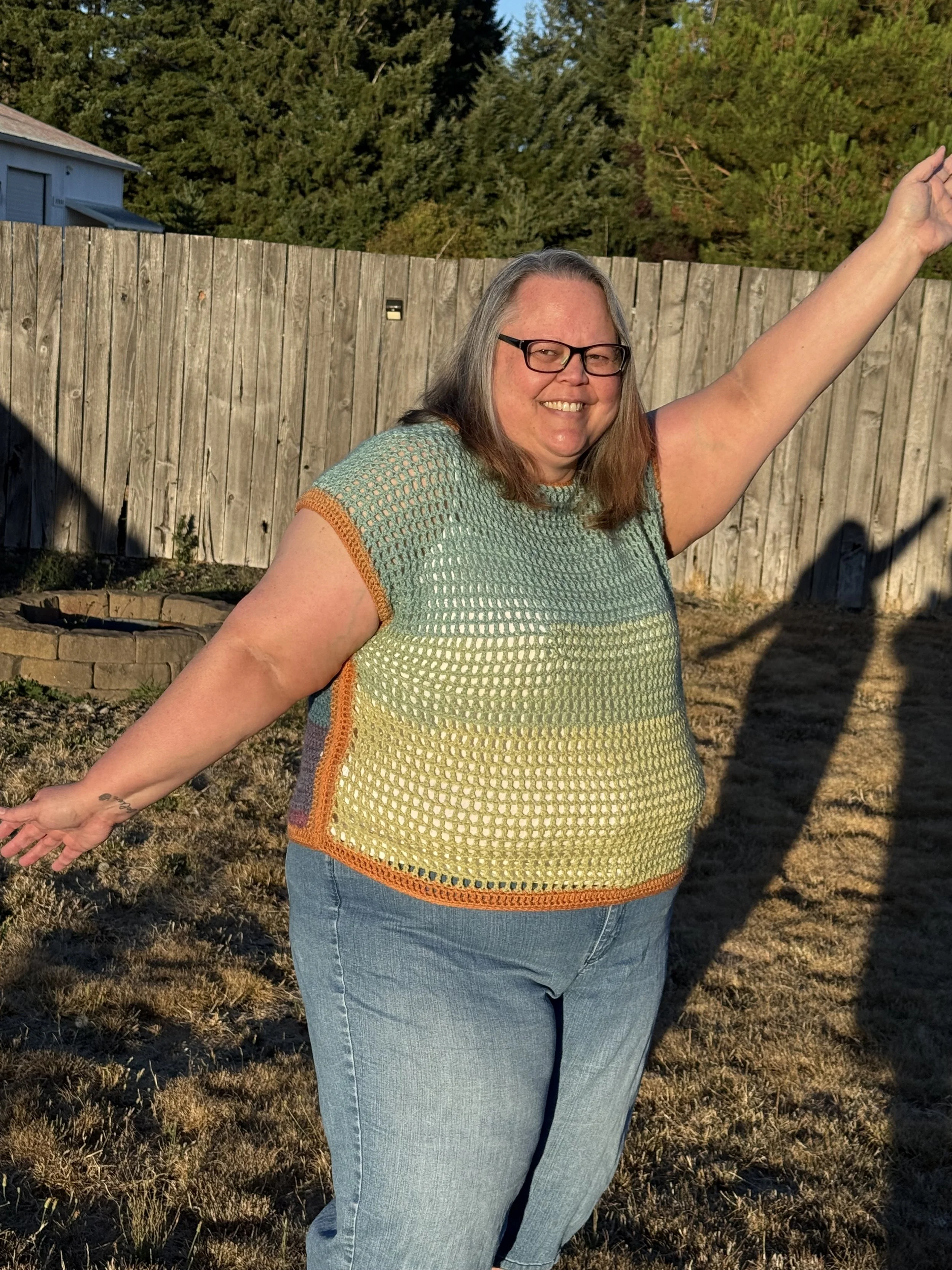 A woman with glasses smiling outdoors near a wooden fence, extending her arms with a cheerful expression.