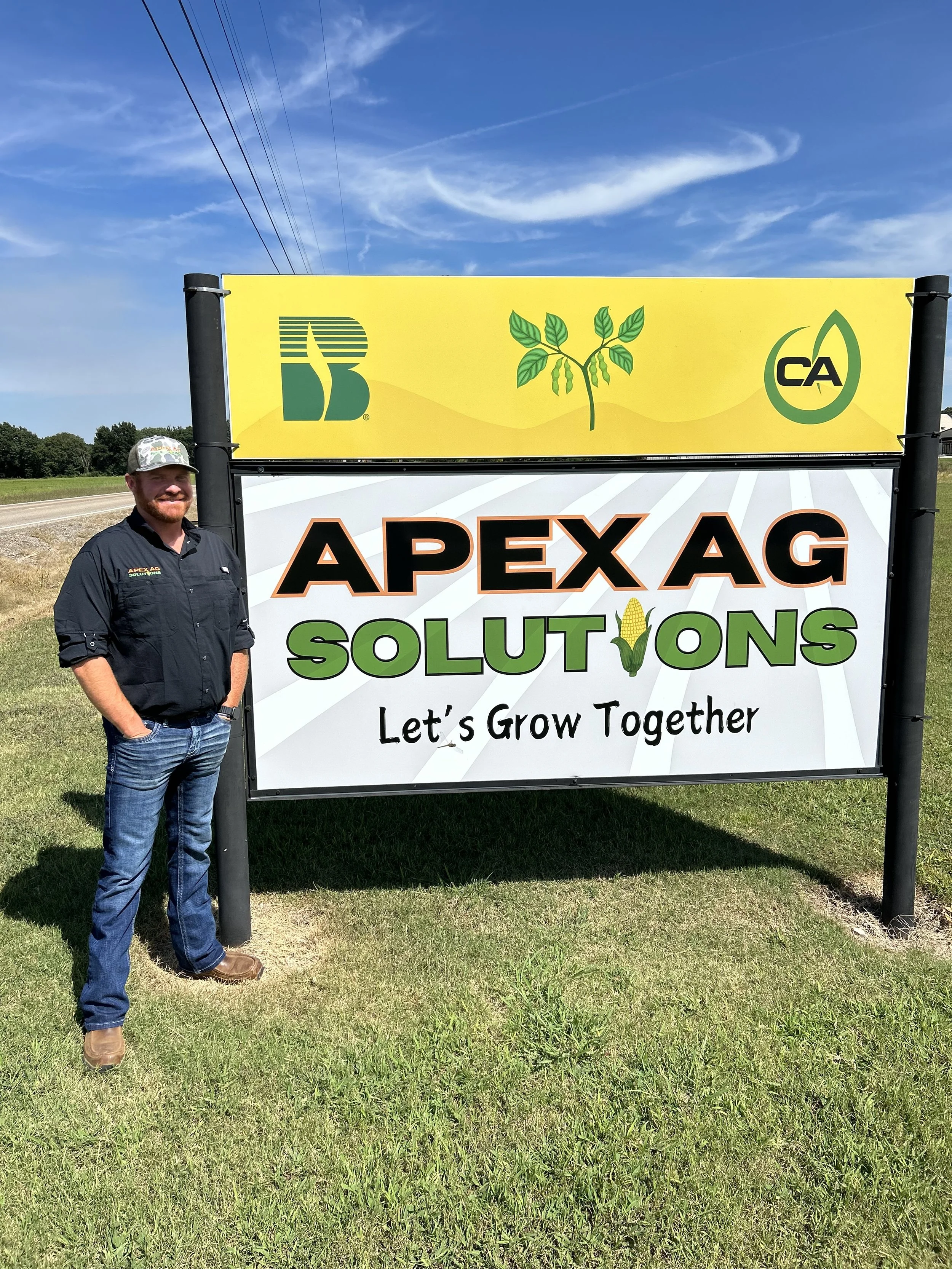 A man standing next to a sign for Apex Ag Solutions on a grassy area near a road, with a clear blue sky overhead.