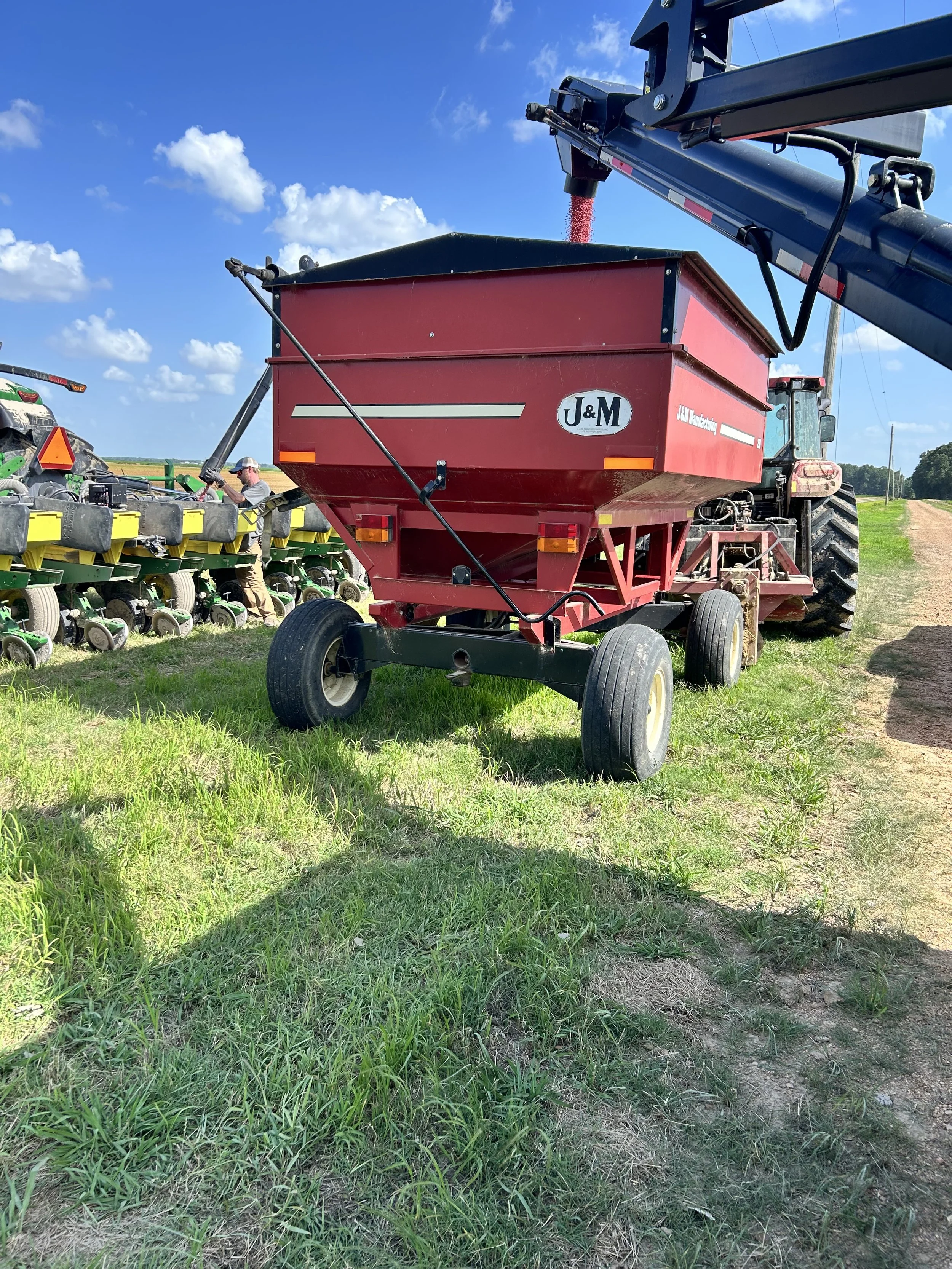 Red agricultural grain cart attached to a tractor, in a field under a blue sky with clouds.