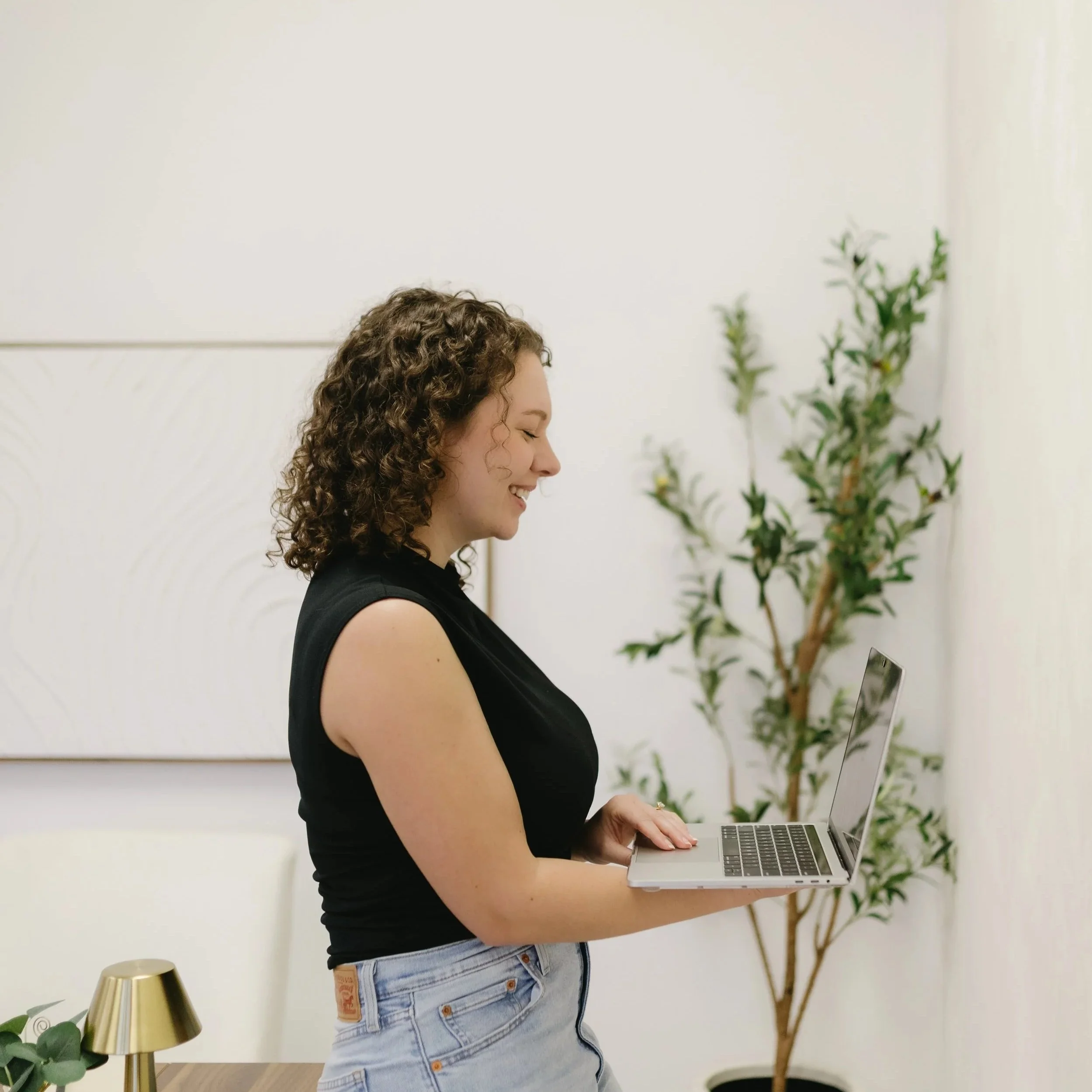 A woman with curly brown hair, wearing a black sleeveless top and ripped jeans, smiling and looking at a laptop she is holding with both hands, standing in a room with a white wall, green plant, and gold lamp.
