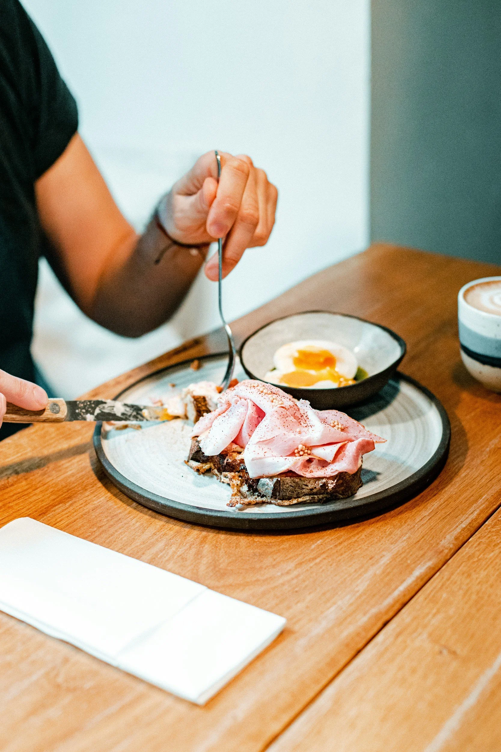 A person holding a fork over a slice of cake with pink frosting and sprinkles on a plate, with a bowl of soft-boiled egg and a cup of coffee nearby on the wooden table.