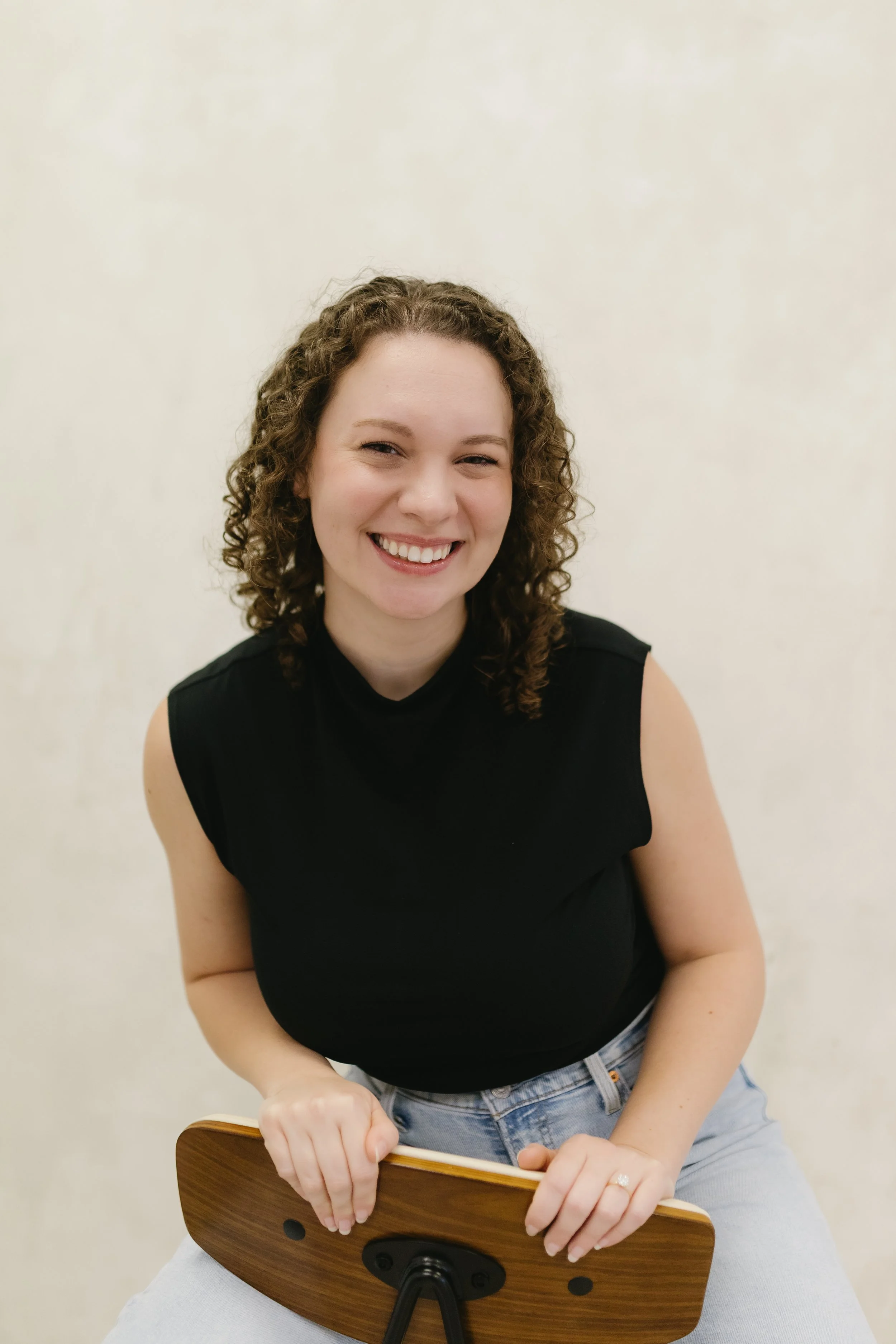 A woman with curly brown hair smiling while sitting on a chair, holding the backrest with both hands, against a plain light-colored wall, wearing a black sleeveless top and light jeans.