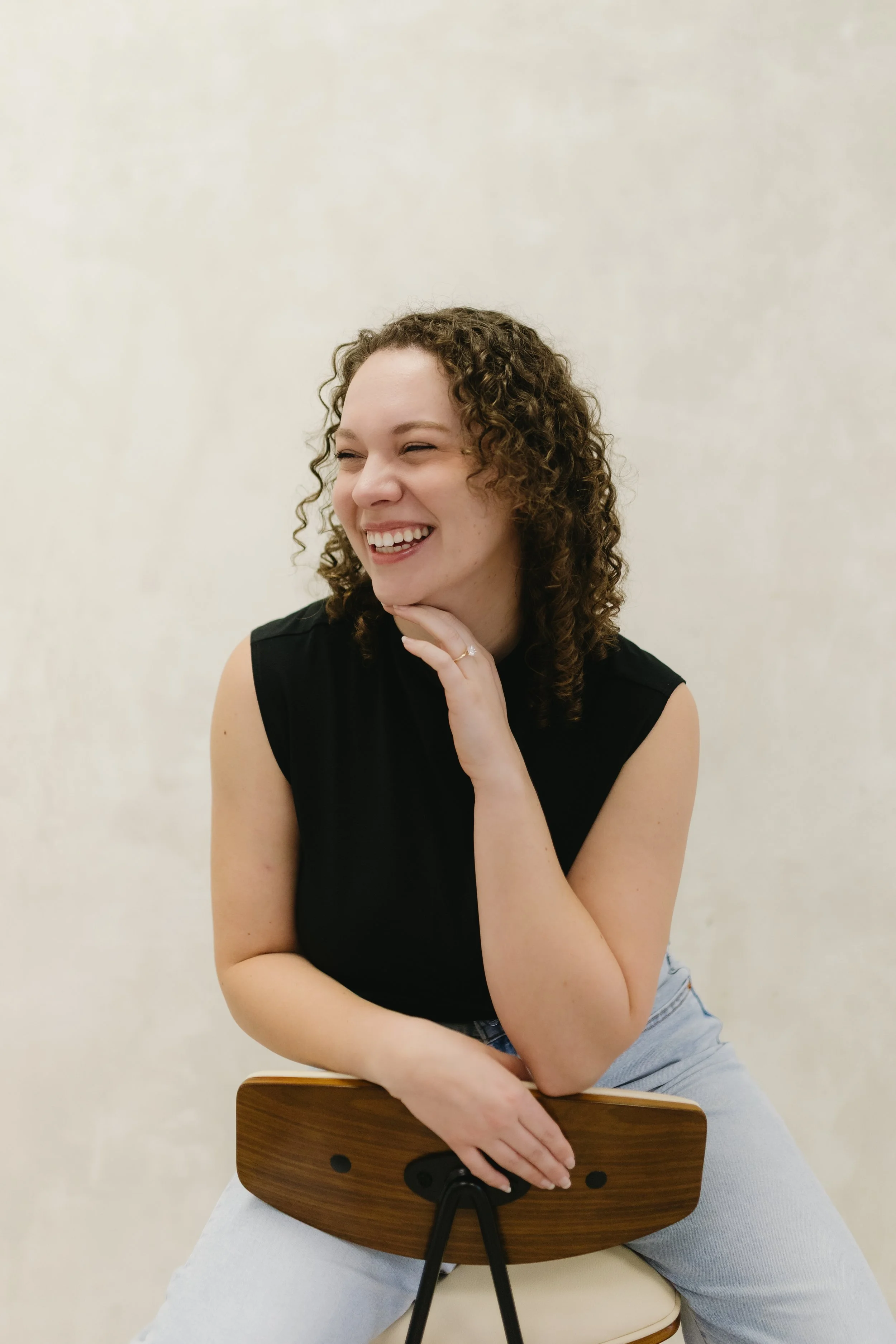 A woman with curly hair smiling and looking to the side, seated in front of a white wall, with one hand resting on her chin and the other on her lap, seen through a camera on a wooden chair.