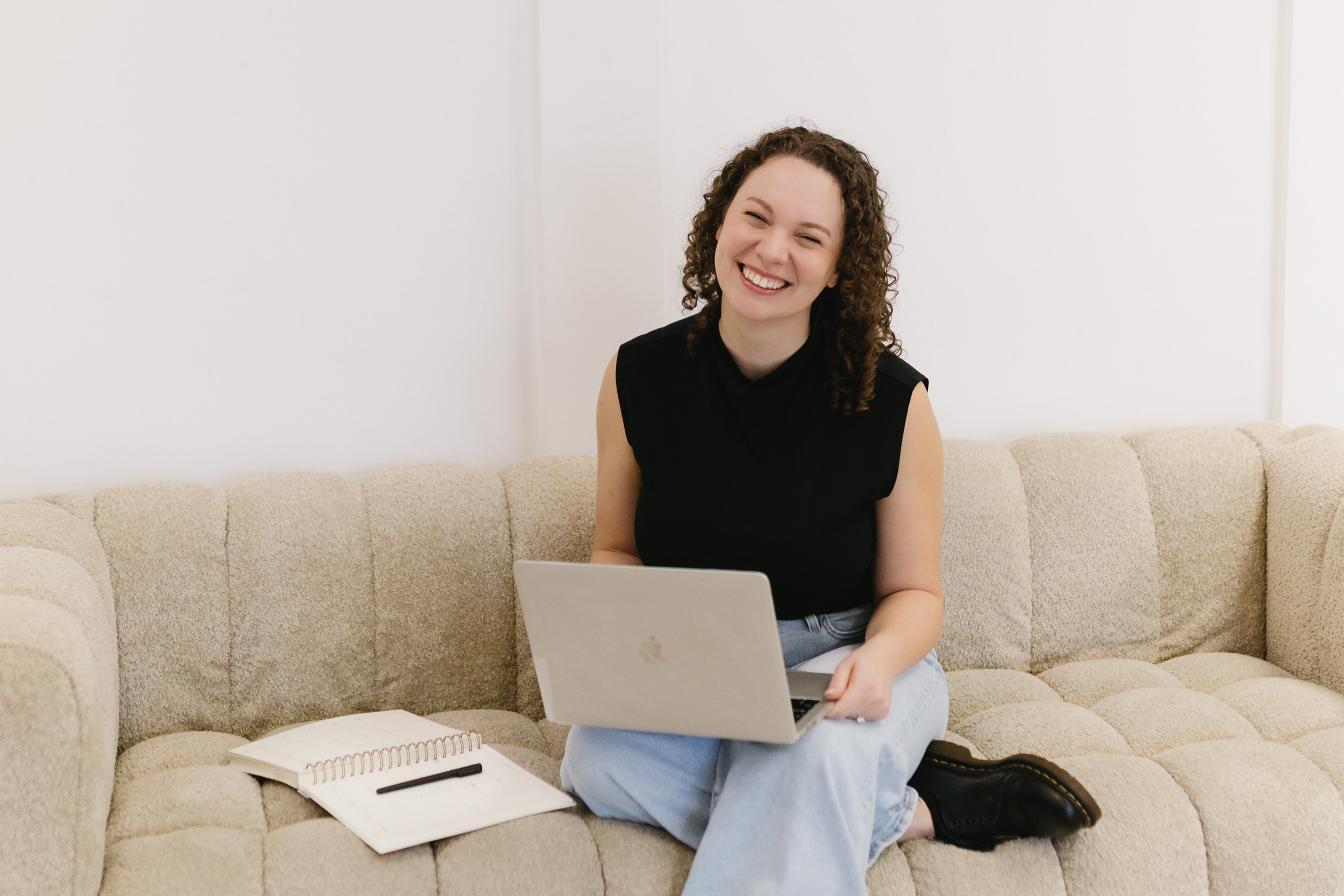 A young woman with curly hair, wearing a black sleeveless top, is sitting on a beige couch, smiling while working on a silver laptop. There is an open notebook and pen in front of her.