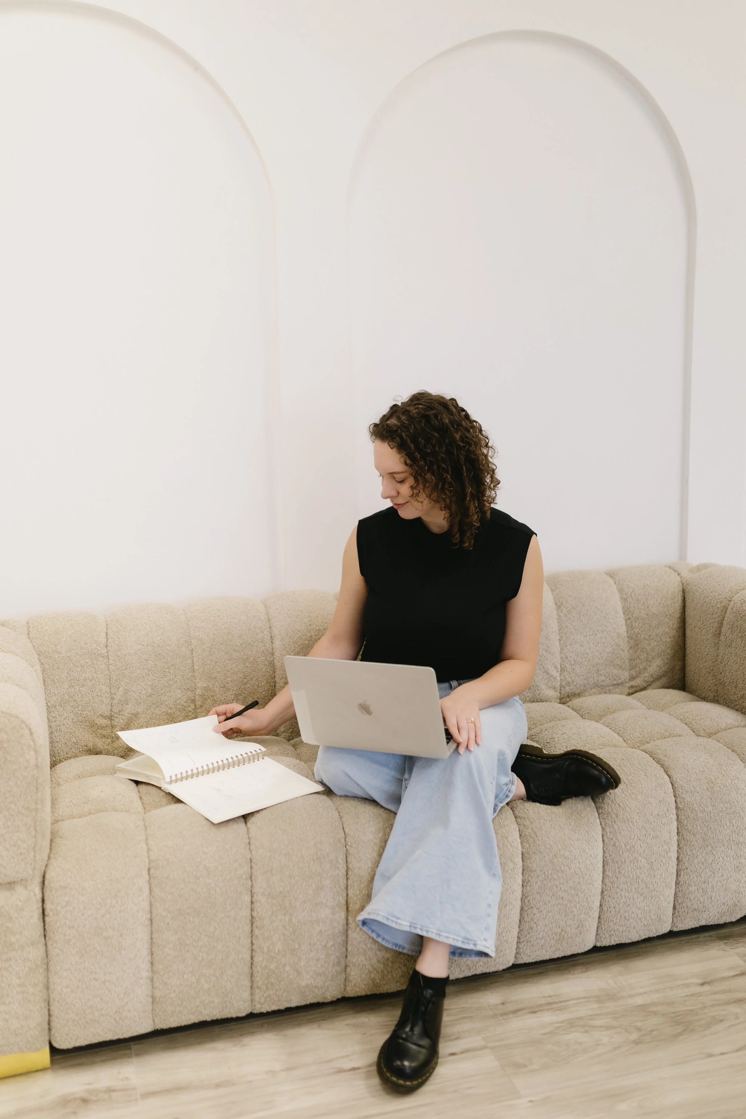 A young woman with curly hair sitting on a beige sectional couch, working with her laptop, writing in a notebook, and surrounded by papers in a room with plain white walls and wooden flooring.