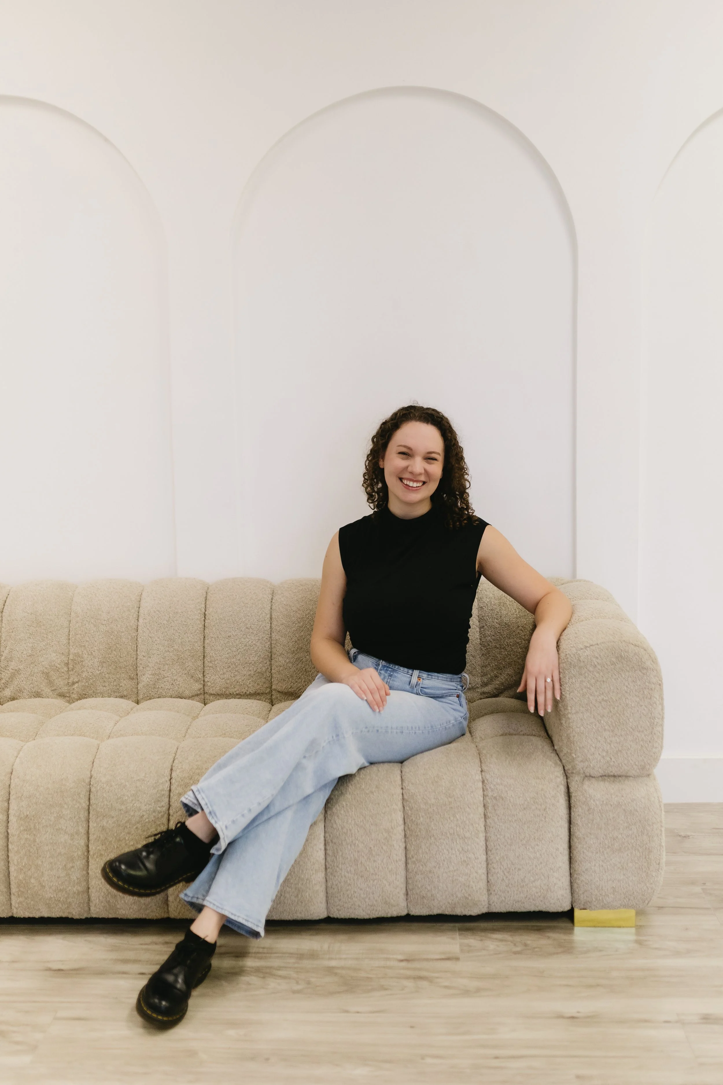 A woman with curly hair sitting on a beige couch, smiling at the camera, wearing a black sleeveless top and light blue jeans, in a room with white walls and light wood flooring.
