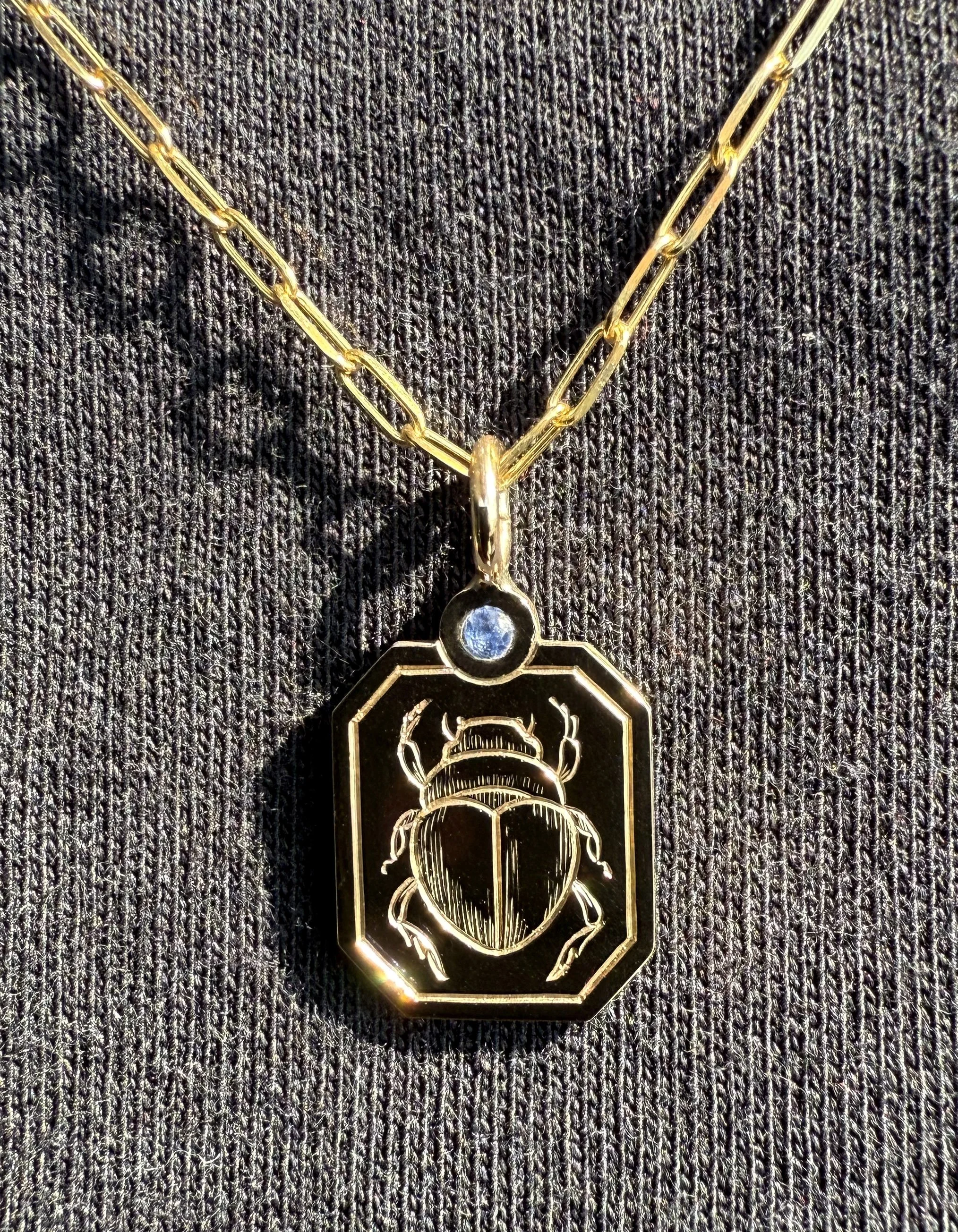 Gold necklace with a pendant featuring an engraved beetle and a small gemstone, placed on dark fabric.