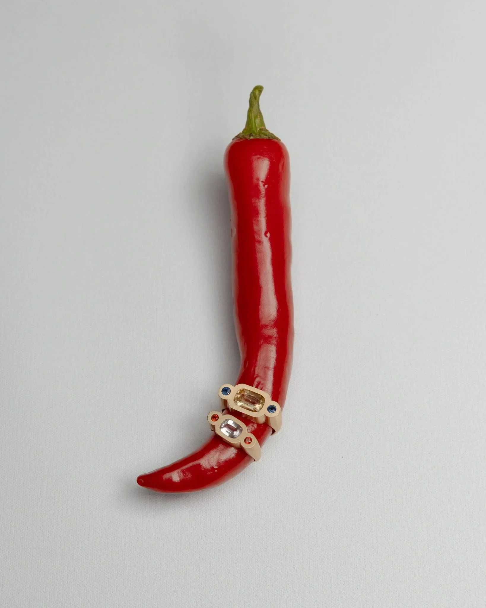 A red chili pepper with two rings placed around its lower part, featuring sapphires and garnets in yellow gold, set against a plain white background. Photographed by Holly Graham