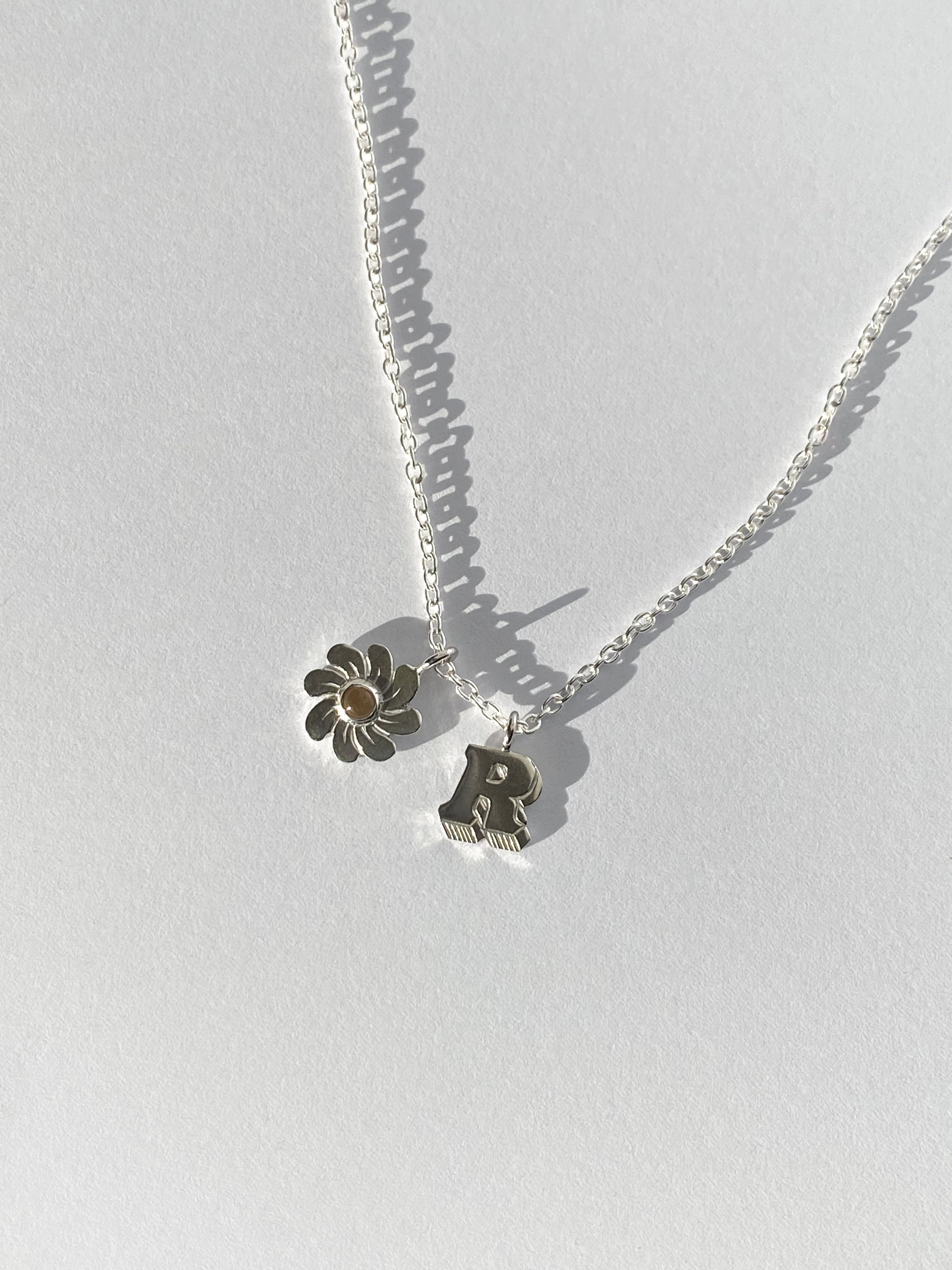 Silver necklace with two charms: a flower with a small circular center and a block letter R pendant. The necklace is on a white background, casting shadows.